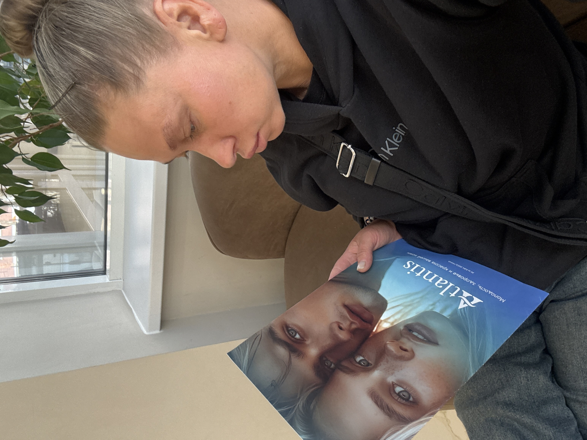 A woman with her hair pulled back into a bun is sitting and looking at a magazine called "Atlantis". She is wearing a black Calvin Klein sweatshirt and gray jeans. A black strap crosses her chest. The woman is sitting near a window with a plant in front of it. The location is most likely in Perm, Russia, as the magazine is written in Russian.