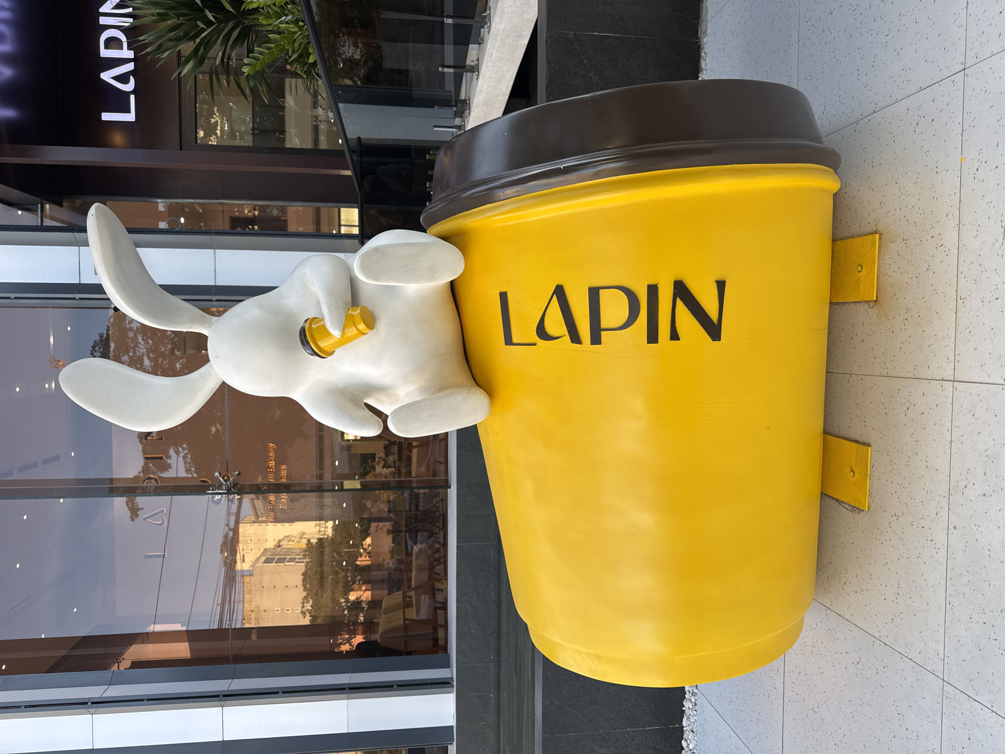 A detailed forensic description of the image is as follows:
The image depicts a whimsical outdoor display in front of what appears to be a café or bakery named "Lapin." The display features a large, bright yellow coffee cup with the word "LAPIN" in bold black letters. The cup has a brown lid and is mounted with yellow brackets to the sidewalk. A white rabbit statue is propped against the coffee cup, holding a golden pipe or straw. The sidewalk is tiled with light-colored, speckled tiles.
Reflections in the windows suggest a city setting with buildings and foliage across the street. The sign "Lapin" is clearly visible at the top of the window display, indicating the location. Given the architectural style and signage, the location is likely in Đà Nẵng, Vietnam.
There are no people visible in the scene.