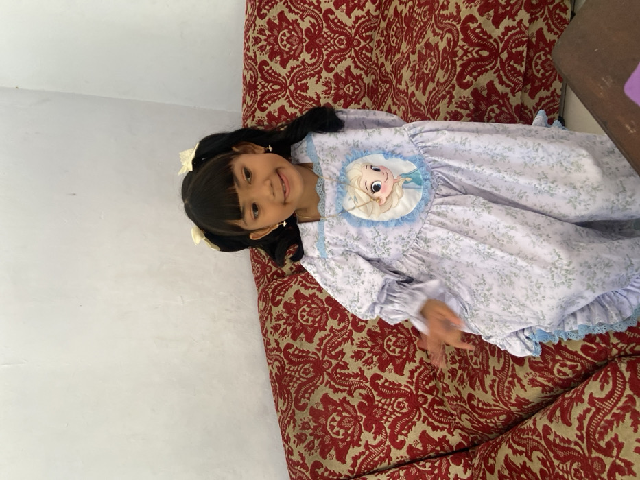 The image displays a young female child seated on a sofa within an indoor environment in Tambakrejo, Indonesia. The child possesses dark hair, styled into two pigtails secured with yellow bows, and straight bangs across her forehead. She wears small earrings and a light lavender dress featuring a subtle green floral pattern, long ruffled sleeves, and an oval appliqué on the chest depicting the cartoon character "Elsa" from Frozen, framed by light blue lace. A thin, gold-colored necklace with a small pendant is visible around her neck, resting over the appliqué. Her mouth is slightly open in a smile, with her tongue partially visible. Her right hand is slightly blurred, indicating movement, positioned away from her body. The child is looking towards the viewer. The sofa is upholstered in a red fabric with a gold or beige damask pattern. Behind the sofa is a plain, off-white wall with minor surface irregularities. A corner of a dark wooden table or furniture item is visible in the lower right foreground. The scene is illuminated by even, natural light.