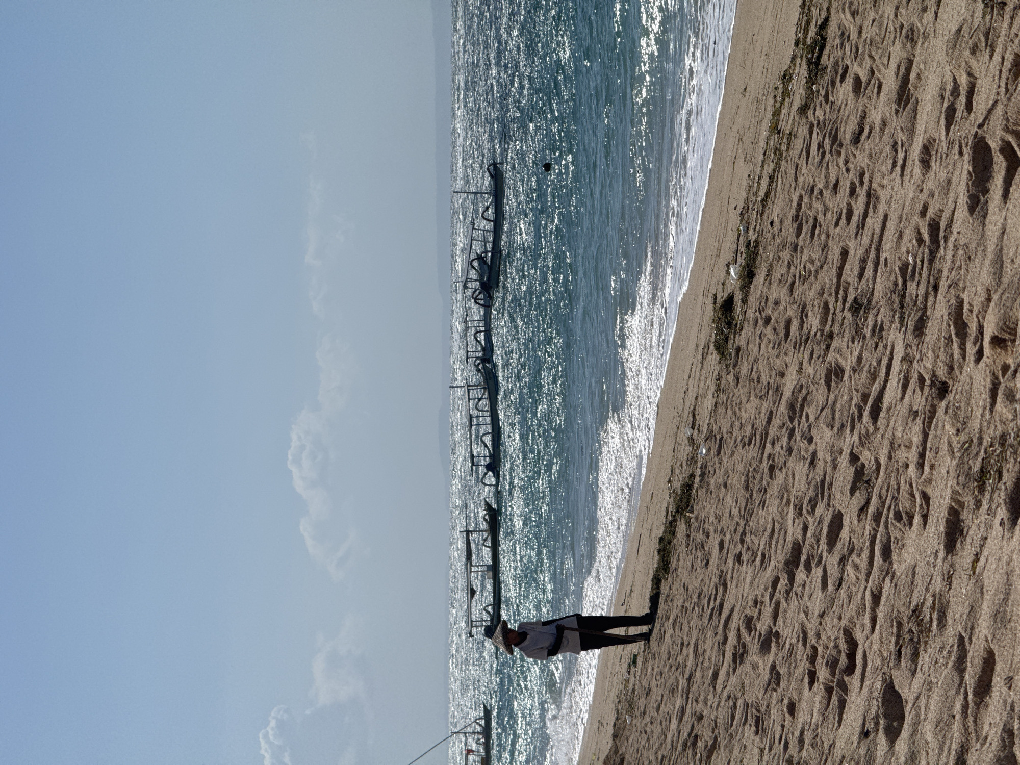 The image depicts a sunlit beach scene, viewed from a slightly elevated position looking down the shoreline. The image, when correctly oriented, shows a wide expanse of sandy beach on the right, the ocean in the middle, and a clear sky on the left.
In the foreground, on the sandy beach, a solitary person stands facing towards the ocean. They are wearing a light-colored long-sleeved shirt, dark trousers, and a wide-brimmed straw hat. They appear to be holding a long, slender pole or tool that extends downwards towards the sand.
The sandy beach is characterized by numerous ripples and textures, likely formed by waves or wind, and shows some patches of dark green vegetation or seaweed near the water's edge.
The ocean, taking up the midground, is a vibrant blue-green, intensely shimmering and sparkling with sunlight reflections across its surface. Gentle white waves can be seen breaking along the shoreline. Extending horizontally into the water from the left side of the image is a dark, low-lying structure, resembling a partial pier or a series of connected posts. A dark, spherical object, possibly a buoy, floats in the water near this structure.
The sky on the left is a clear, light blue, with a few soft, white clouds visible towards the bottom left. The distant horizon line beyond the water is hazy, suggesting atmospheric conditions or distant land.
The scene conveys a bright, sunny daytime setting. There is no visible text in the image.