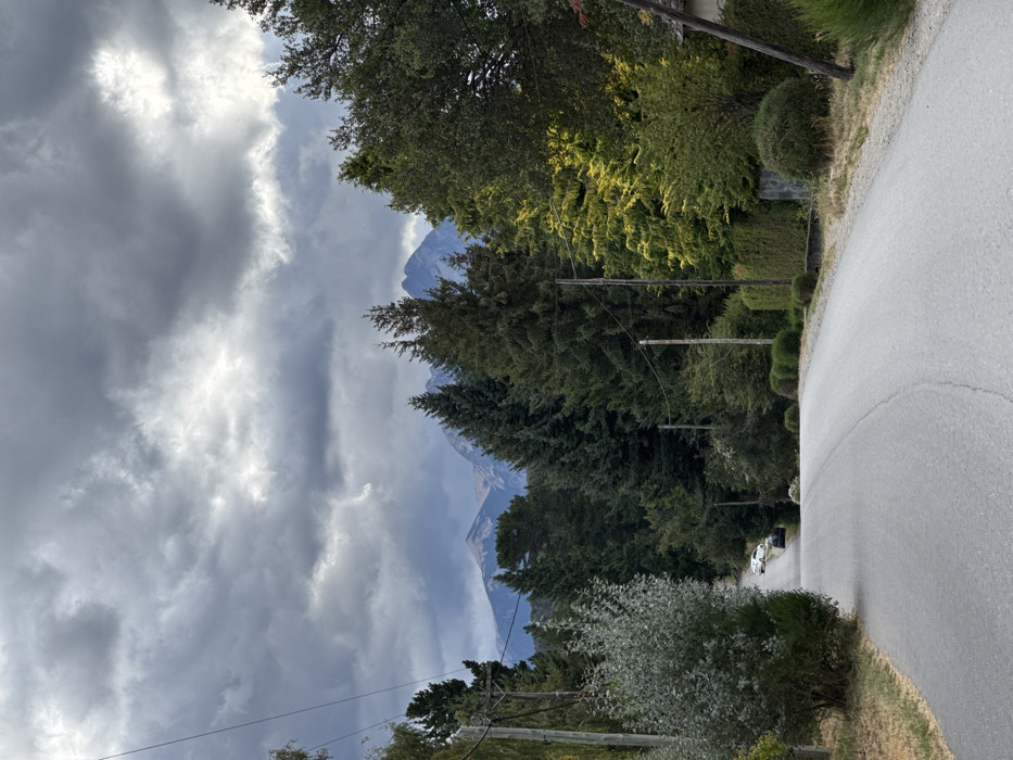 A winding road curves through a green landscape. Trees line the road, and beyond them, a mountain range is visible in the distance under a cloudy sky. Telephone poles run alongside the road, emphasizing the rural setting. A vehicle is parked further down the road. The overall scene evokes a sense of peacefulness and natural beauty.