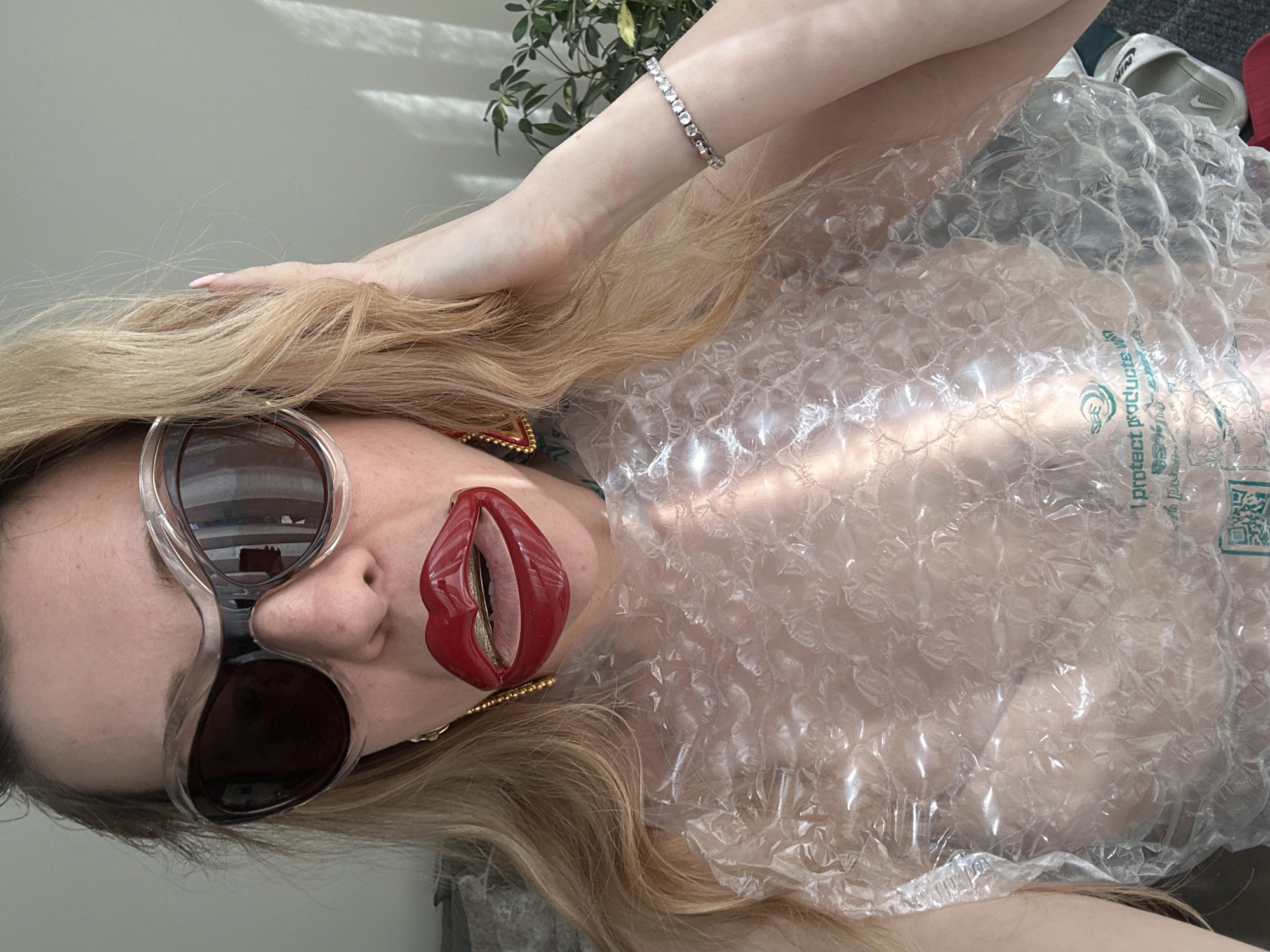The image displays a person, likely female, positioned horizontally with her head towards the left side of the frame. She has long, wavy blonde hair flowing over her shoulders and across the upper part of the image. Her face features large, clear-framed sunglasses with dark lenses, obscuring her eyes. Her lips are adorned with bold red lipstick, and she wears a distinctive red, lip-shaped earring on her left ear. A silver tennis bracelet is visible on her raised left arm, with her hand resting in her hair near the top of the composition.
The person's body is largely covered in clear plastic bubble wrap, which functions as an unconventional garment. The bubble wrap reflects bright natural light, creating numerous highlights and shadows. Green text is printed on the bubble wrap, explicitly stating "i protect products" alongside a QR code and other smaller, possibly foreign, text.
In the background, a plain light grey wall is visible behind her head, featuring distinct shadows cast by a window blind. Towards the upper right, a green leafy houseplant adds an organic element. Further to the right, in the background, light-colored footwear, including what appears to be a white Nike slide, can be faintly seen. The strong, direct light suggests a daytime setting, and the overall presentation implies an artistic, conceptual, or fashion-oriented photoshoot.