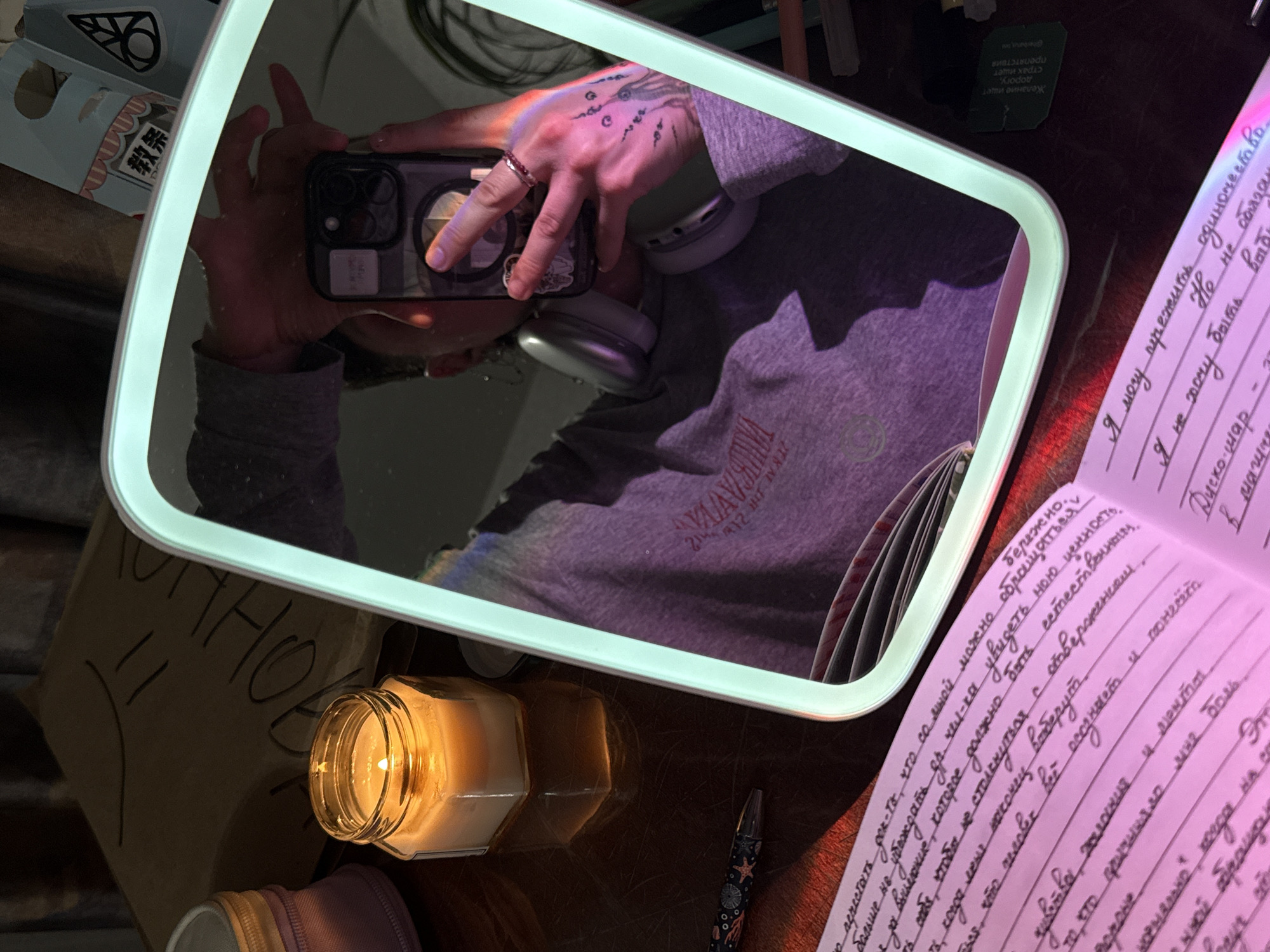 The image shows a person taking a selfie in a lighted mirror. The person is wearing a gray sweatshirt, has tattoos on their hand, and is wearing headphones. In the reflection, the phone's camera array is visible. Around the mirror are various items, including a lit candle in a glass jar, a notebook with handwritten text in Russian, a pen, and miscellaneous objects. The writing appears to be personal reflections. The environment suggests an indoor setting, possibly a home study or creative workspace. The presence of Russian text indicates the location is likely in Russia, potentially Saint Petersburg given the prevalence of such imagery from that city online.