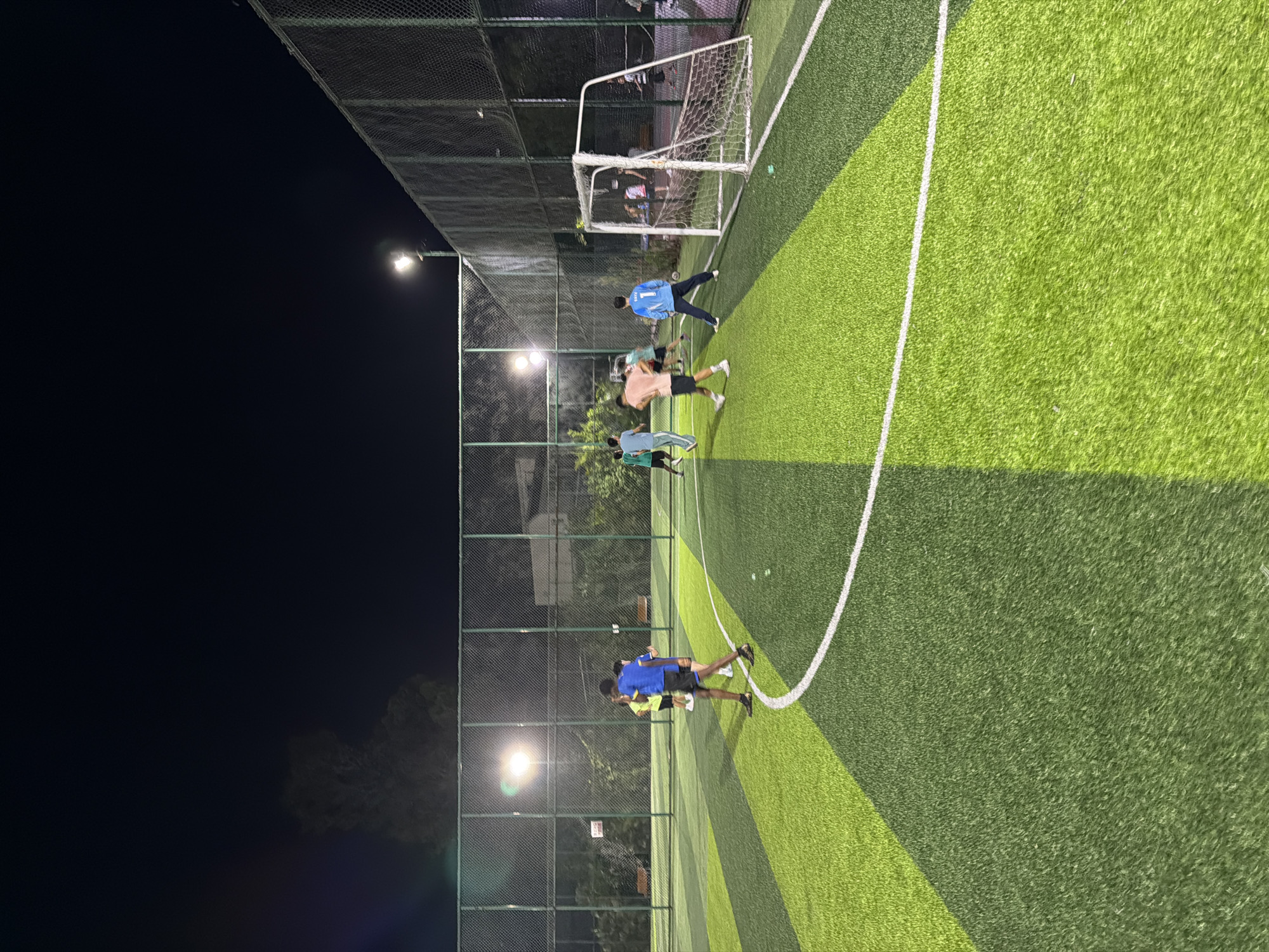 At night, a group of people are playing soccer on an artificial turf field. The field is illuminated by overhead lights and has distinct green and lime green sections separated by white lines. A white soccer goal with a net is visible on the right side of the frame. Several individuals are actively engaged in the game, some running with the ball and others positioned on the field. The background shows a chain-link fence, trees, and some buildings, suggesting an urban or suburban setting. The scene appears to be in the city of Jiangjin, China.