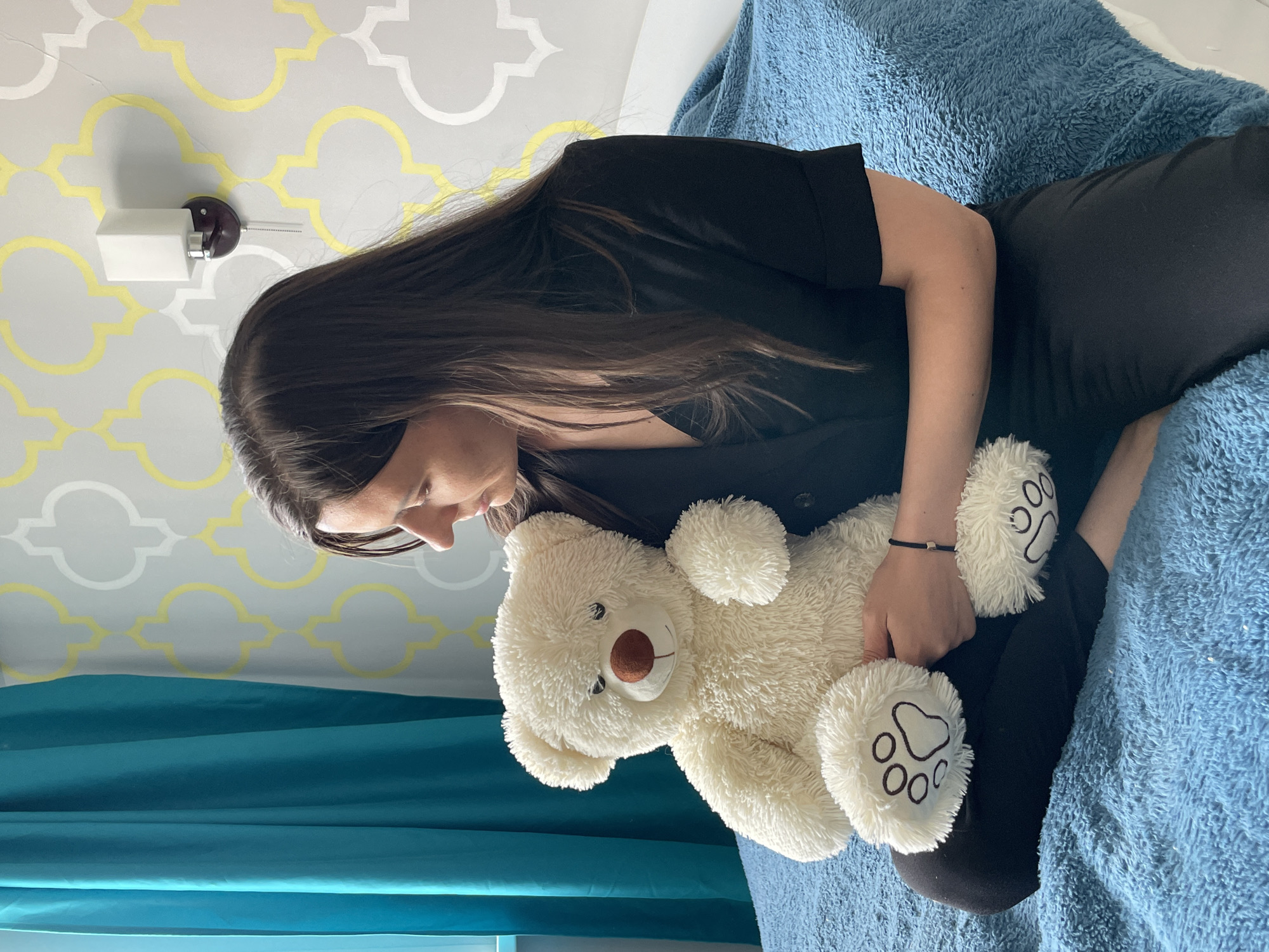 A person with long, dark hair is sitting on a blue, textured surface, possibly a bed or couch, and hugging a cream-colored teddy bear with paw prints on its feet. The person is wearing a black top and dark pants. Behind them, a wall is visible with a gray and yellow patterned wallpaper and a white wall sconce. A blue curtain or sheet partially obscures the lower left corner of the image. The location is Unknown, Unknown.