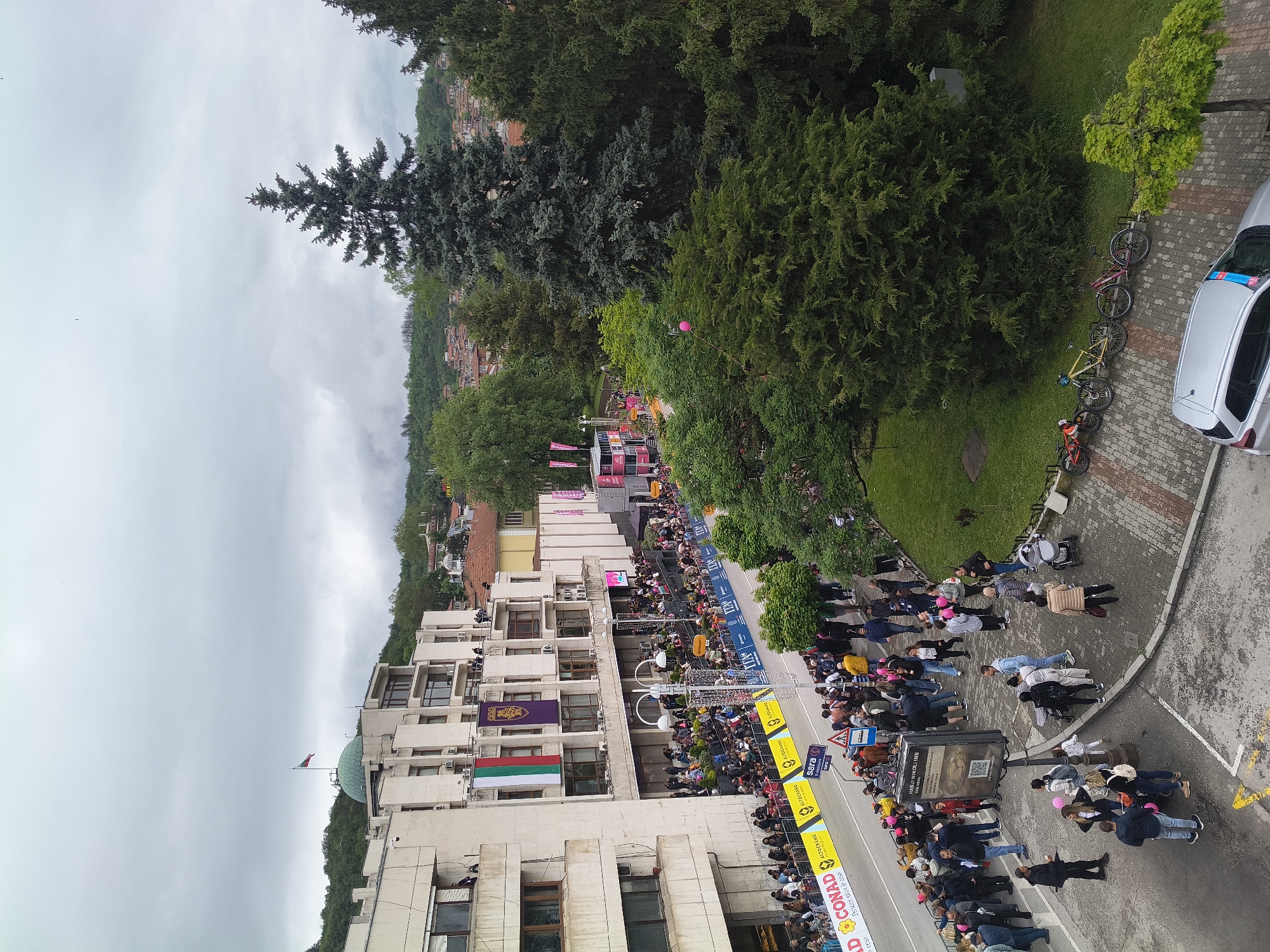 An aerial perspective reveals a scene in Veliko Tarnovo, Bulgaria. A crowd of people lines the street, observing an event. Banners and barricades suggest an organized gathering, possibly a race or parade. Buildings with Bulgarian flags frame the street. Trees and foliage create a green backdrop against a cloudy sky. A parked car and bicycles rest nearby. Pedestrians gather on a paved area to observe the event. The environment is urban, with a mix of nature and constructed elements.