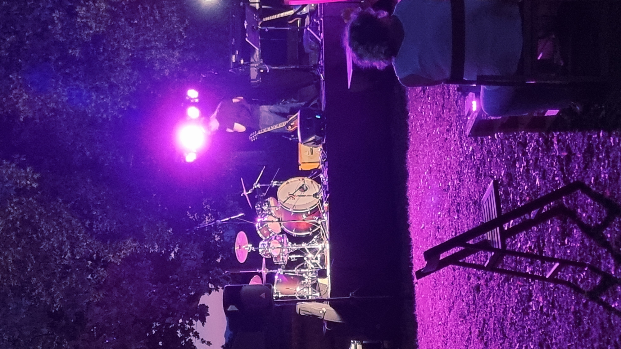 The image depicts an outdoor evening concert scene, likely in the municipality of Sant Joan Les Fonts, Spain, illuminated by vibrant purple stage lighting. In the foreground, a person with curly hair is seated, viewed from behind, facing the stage. To the left, a folded dark wooden chair sits on a gravelly surface. The stage, covered with a dark cloth, features a drum set on the left, with various cymbals and drums. A male figure, wearing a dark t-shirt and jeans, stands near the center of the stage, facing away from the camera, with a guitar leaning against a speaker. Other musical instruments, including what appears to be a keyboard and another guitar, are also visible on the stage. The background is dominated by the dark silhouettes of trees, their leaves illuminated by the purple light. The overall atmosphere suggests a performance taking place in a natural or park-like setting.