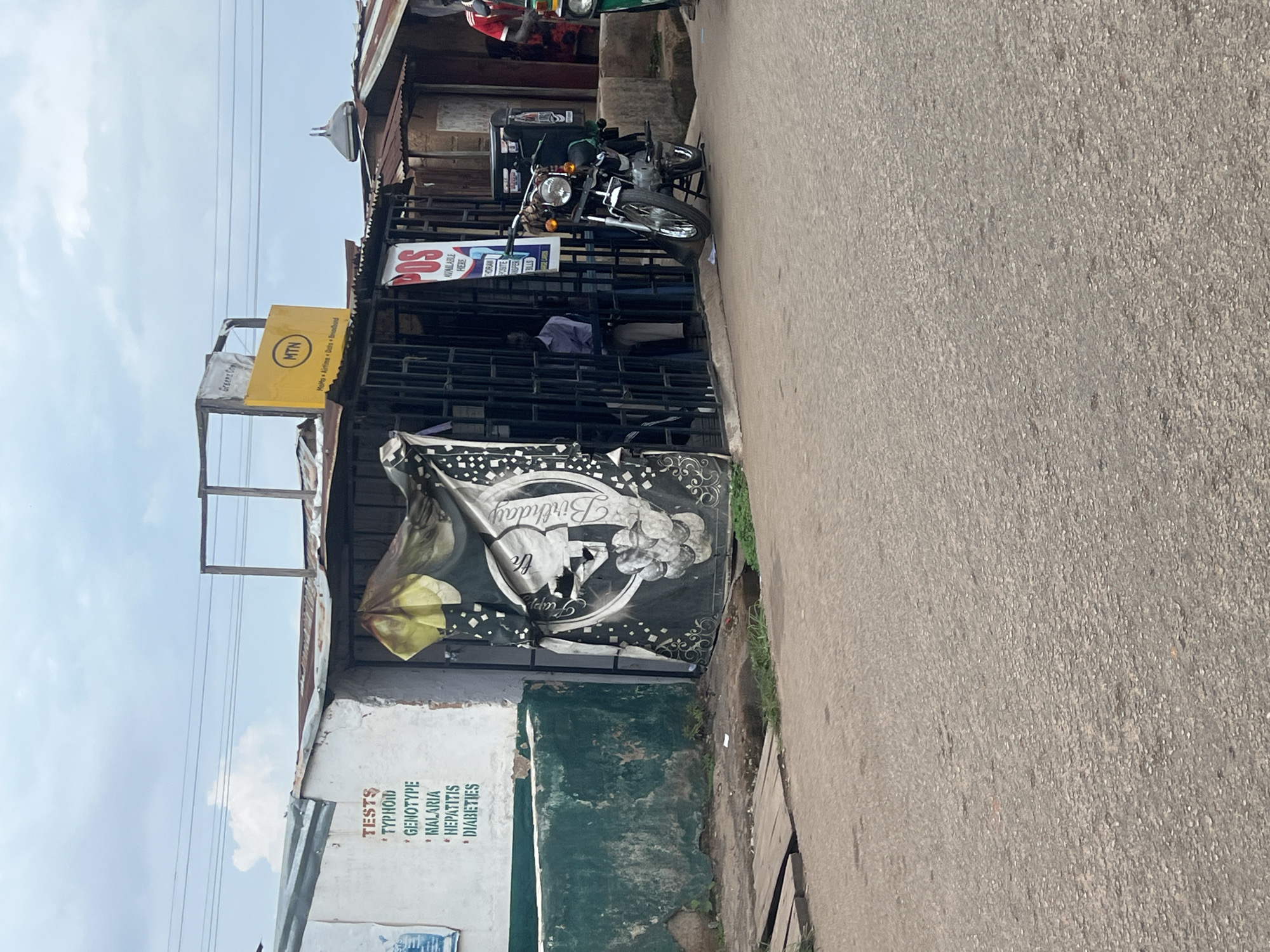 This is a forensic, matter-of-fact description of a scene in Jos, Nigeria. The image depicts a street scene with a small building complex. On the left side, a white wall with green paint at the bottom has text indicating "TESTS" with a list including "TYPHOID", "GENOTYPE", "MALARIA", "HEPATITIS", and "DIABETIES". To the right of this wall, an establishment with a metal grate entrance is visible. A dark banner with a "Happy Birthday" message and an image of a woman is partially covering the entrance. Above this establishment, a yellow sign with the MTN logo and text "MoHtbq Airtime Data Broadband" is mounted. Further to the right, another small building with a rusted corrugated iron roof is present. A motorcycle with a black storage box is parked in front of the MTN establishment, and a person in a blue shirt and grey trousers is visible inside the establishment. On the far right, a person in a red shirt is standing next to a green tricycle. The ground in front of the buildings is a paved road. The sky is overcast with scattered clouds.