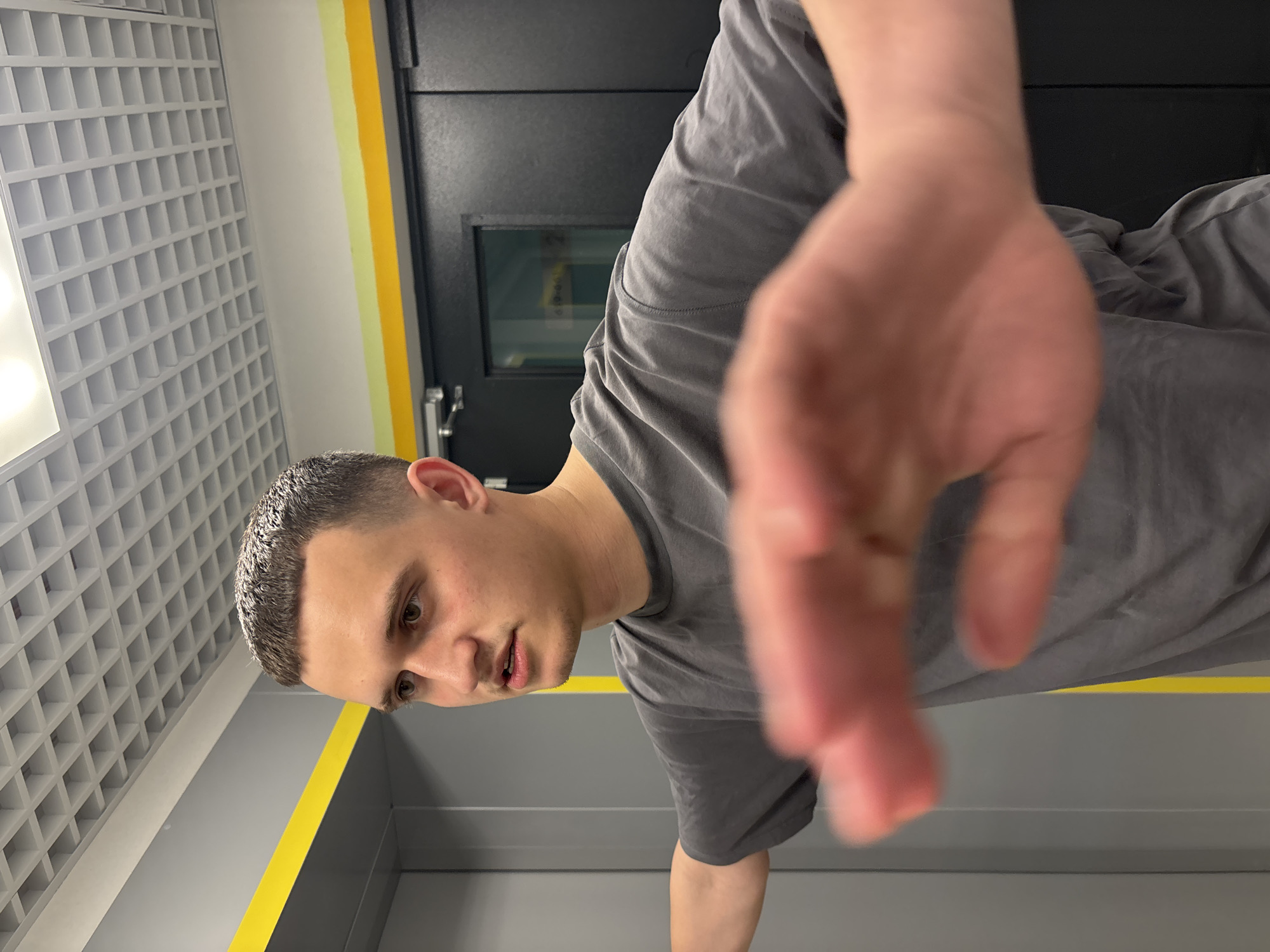A man is facing the camera with his arm outstretched. He has short, dark hair and is wearing a gray t-shirt. The background includes a gray wall with a yellow stripe. There is a grey wall with a grid pattern. A black door is also visible. There is no evidence to indicate that this image was taken in the city of Vsevolozhsk, Russia.