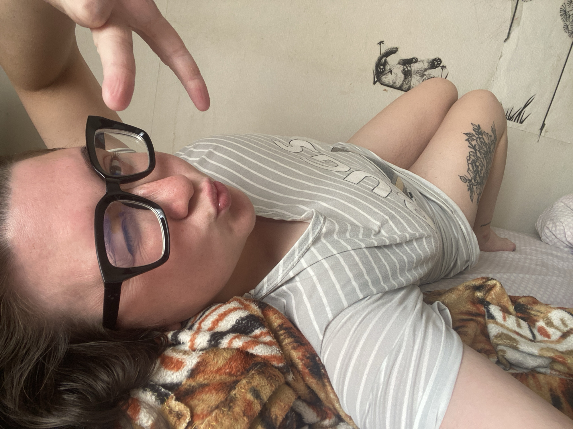 The image captures a person lying on their side in an indoor setting, likely a bedroom, with a casual and relaxed atmosphere. The person, appearing to be female, is positioned horizontally in the frame, looking towards the viewer with a slight head tilt. She wears black, cat-eye style eyeglasses, which rest slightly askew on her face, revealing her left eye. Her mouth is slightly open, suggesting a pout or relaxed expression.
She is dressed in a light grey and white striped top, with partial text visible on the front, including the word "LUCKY" and other indecipherable letters. A patterned blanket, possibly featuring a tiger or animal print in shades of brown, orange, and black, partially covers her body and the bed she is on.
One of her hands is raised and bent, making a gesture in the upper left of the frame. Her legs are bent at the knees, and a detailed tattoo of floral or botanical design is visible on her right thigh. The background consists of a plain, light-colored wall. Two simple drawings or posters are affixed to the wall: one depicts a cat lying on its back, and another shows vertical lines that resemble dandelions or similar plants.
The scene suggests a moment of rest or relaxation in a home environment. While the location context is Chaikovsky, Russia, no specific geographical cues are visible within the image itself. The lighting is ambient, without indications of time of day or weather.