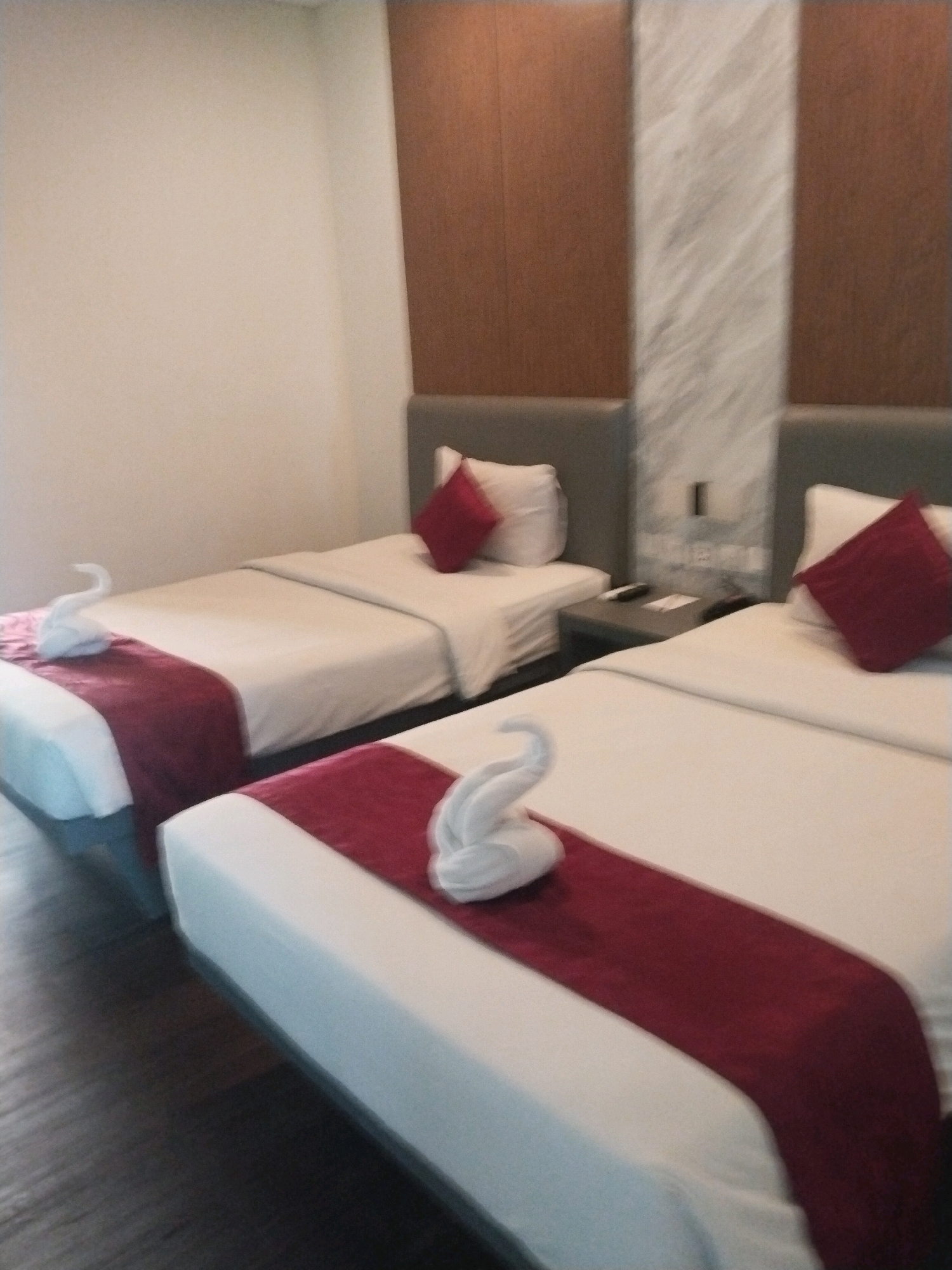 The image shows two beds in a hotel room. The beds are made with white sheets and have a maroon runner across the foot of each bed. There are red square pillows on each bed. On the runner of the bed in the foreground, there is a white towel folded into the shape of a swan. On the bed in the background, there is also a white towel folded into the shape of a swan. To the right of the bed in the foreground, there is a small table with a phone and a remote control on it. The walls are a light color, and there is a wooden panel behind the beds. The floor is made of dark wood. The lighting is soft and even, suggesting it is daytime or a well-lit interior. This appears to be a standard hotel room in a city like Depok, Indonesia, offering comfortable lodging for travelers.