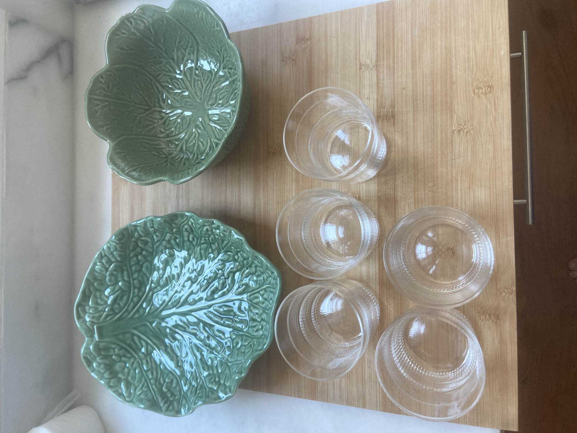 This media file shows two green ceramic serving dishes shaped like cabbage leaves, placed on a light-colored marble countertop. To the right of the dishes, on a wooden cutting board, are five clear glass cups. The cutting board has a metal handle on its right side, suggesting it's part of a kitchen island or drawer. The scene is indoors, likely a kitchen, with soft lighting. No people are clearly visible, though a blurry foot appears in the lower right corner. There is no visible text in the image. The overall impression is one of domestic preparation or presentation of food or drinks. The location context of Matosinhos, Portugal, does not provide any specific visual cues within the image itself.