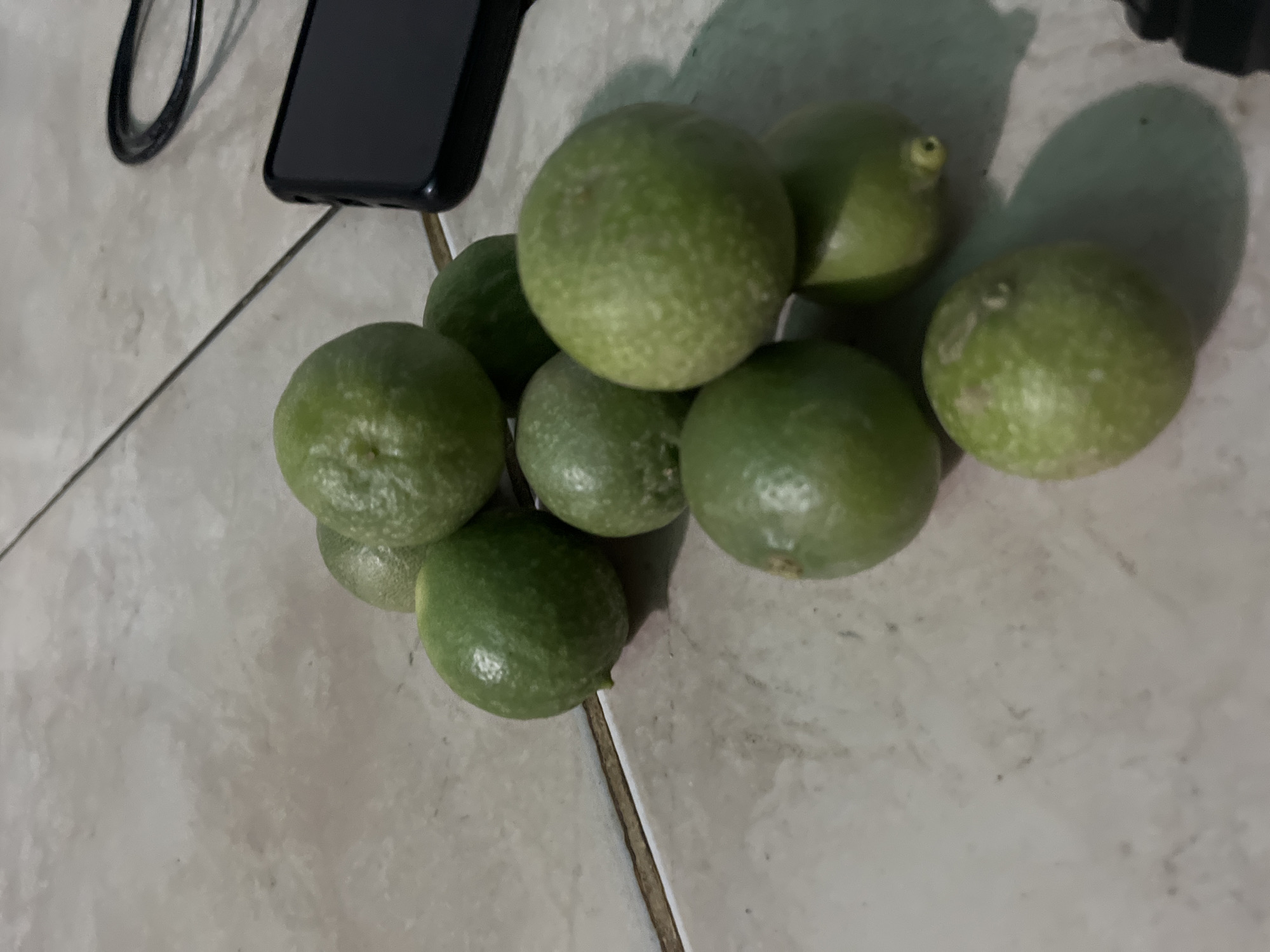 A close-up shot depicts a collection of approximately nine limes on a tiled surface. A black smartphone and a portion of a dark cable are visible in the upper left corner. The image was likely taken indoors, given the tiled floor and ambient lighting. There's no information to suggest the location is in the city of Juan Griego, Venezuela.