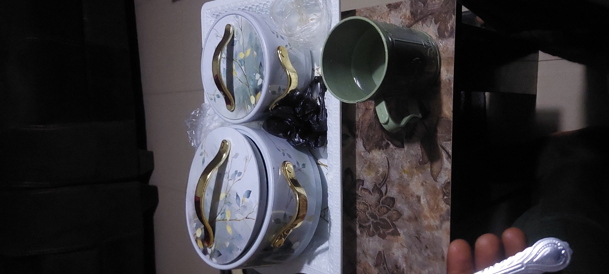 In an indoor setting, two serving dishes with floral patterns and gold handles rest on a white tray. To the right, a green mug with a handle sits on a textured surface, filled with a light-colored liquid. A hand holding a spoon is visible in the lower left corner. The background appears to be a dark, indistinct surface. The scene is likely in a home or dining area.