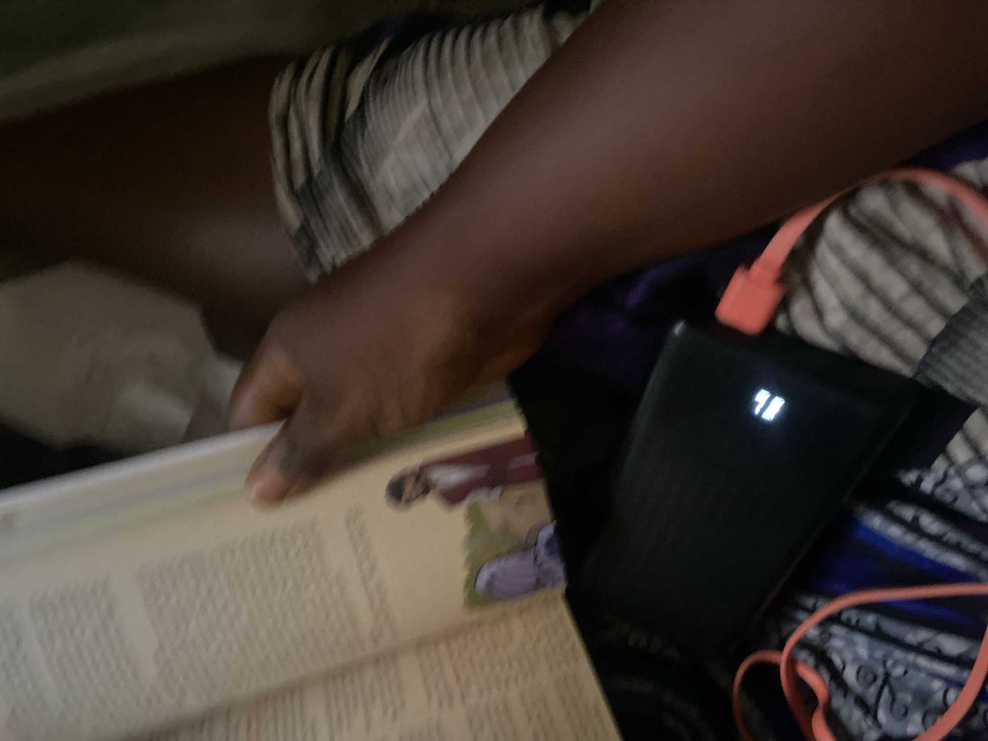 The image depicts a close-up view of a person's arm and legs, as well as an open book and a power bank. The person's skin tone is dark, and they are wearing striped fabric on their legs. The book appears to be a religious text, possibly the Bible, with an illustration of a man in red standing next to a purple figure. A black power bank is placed on a patterned surface, and an orange cord is connected to it. The lighting suggests an indoor setting. The image does not contain any information that would allow for the determination of the location being in the city of Jauro Yinu, Nigeria.