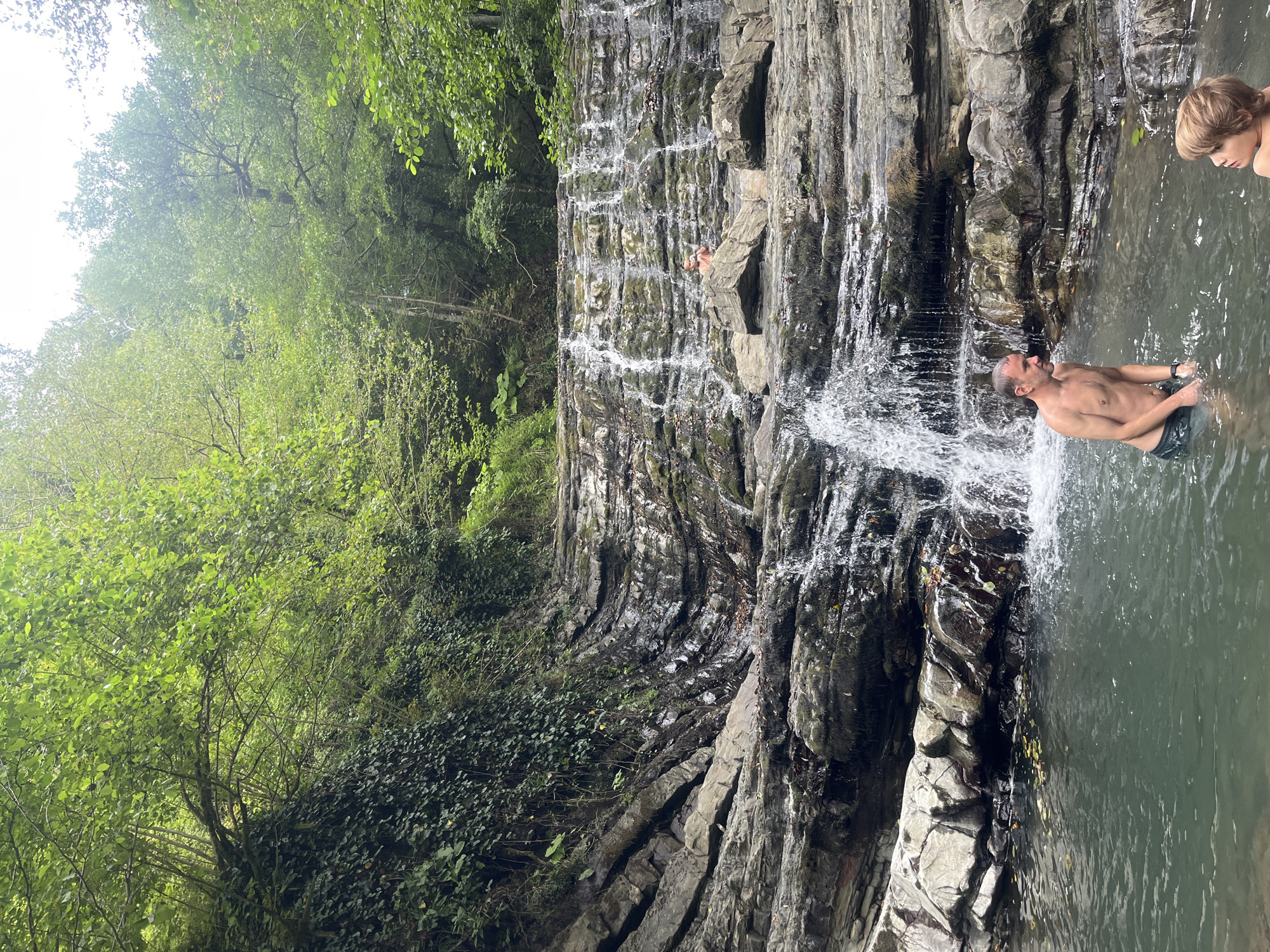 The image shows a natural waterfall cascading over layered rock formations in a lush, green forest setting. In the foreground, a man and a young boy are standing in a pool of water at the base of the waterfall. The man, shirtless and wearing swim trunks, is facing to the right. The boy, with blond hair, is facing to the right and looking up towards the man. The water is clear and reflects the surrounding greenery. The waterfall itself is multi-tiered, with water flowing over numerous rocky ledges. The rocks are wet and appear to be covered in moss and vegetation in places. The background is filled with dense trees and foliage, indicating a natural, undeveloped environment. This scene is located in the Khostinskiy district of Sochi, Russia, a region known for its natural beauty and numerous waterfalls.