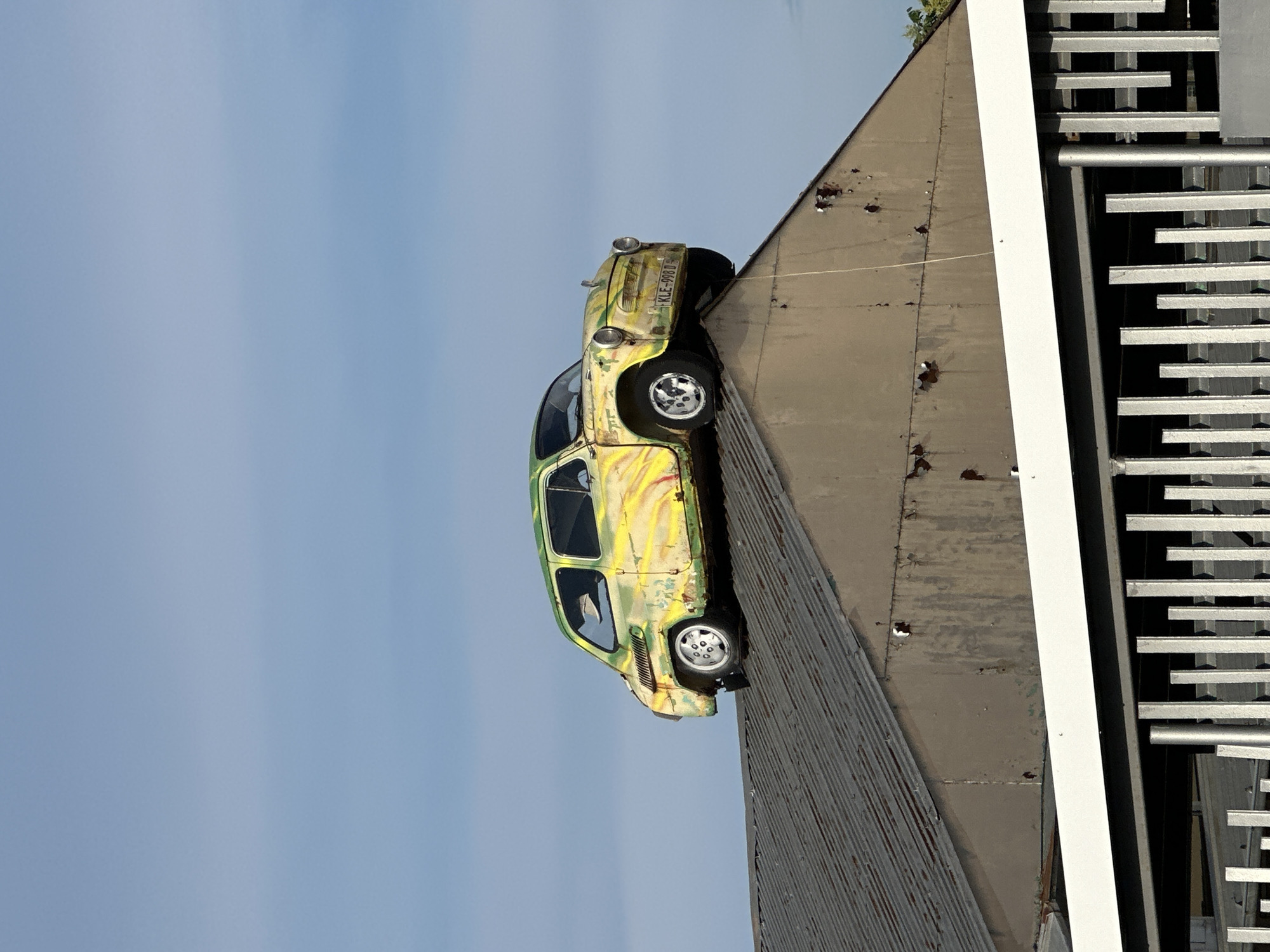 An abandoned, yellow and green vintage Fiat 600 is perched atop the pointed roof of a weathered, corrugated metal industrial building. The car exhibits signs of decay, with peeling paint and rust. The building's facade is also deteriorating, showing rust spots and worn siding. In the foreground, a modern metal fence with vertical slats creates a stark contrast with the older structures. The sky is a clear, pale blue with faint clouds, suggesting daytime. The location is identified as Lyaskovets, Bulgaria.