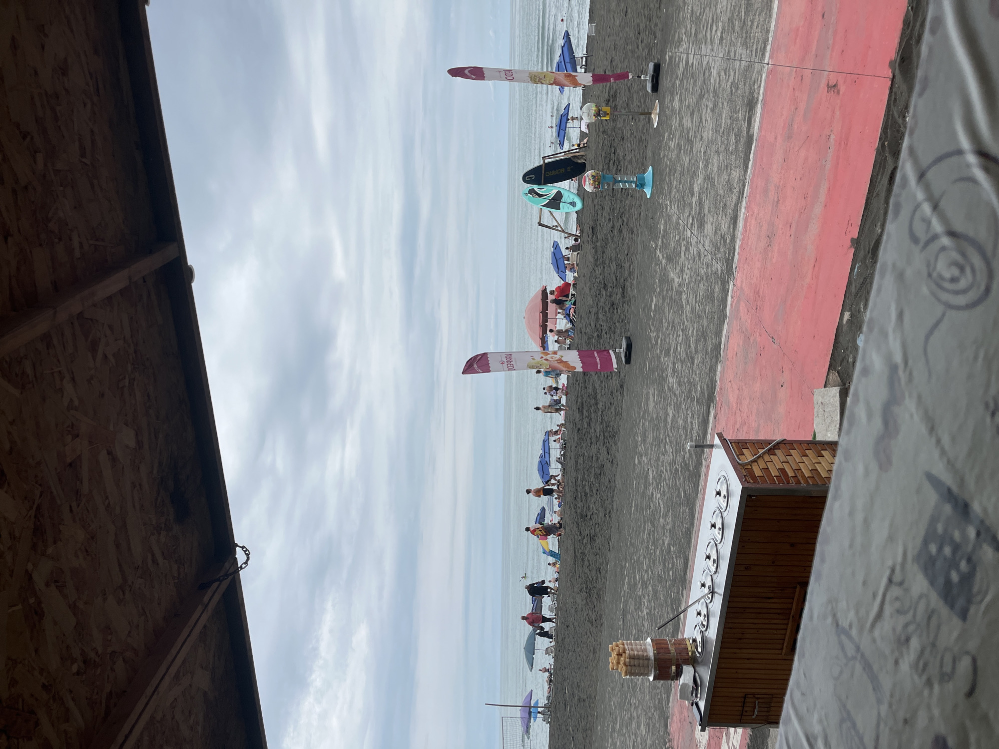 The image depicts a cloudy day at Ureki, Georgia's Black Sea coast, a beach known for its magnetic sand. In the foreground, a wooden ice cream cart with metal containers is visible, topped with a stack of ice cream cones. A red-painted walkway runs parallel to the beach. The beach itself is covered in dark sand, with numerous beachgoers, umbrellas, and surfboards scattered across it. The sea stretches to the horizon under a gray, overcast sky. Beach flags, gumball machines, and a volleyball net are also present on the sand. The image appears to be taken from a covered structure, possibly a restaurant or cafe, with exposed wooden ceiling panels and beams.