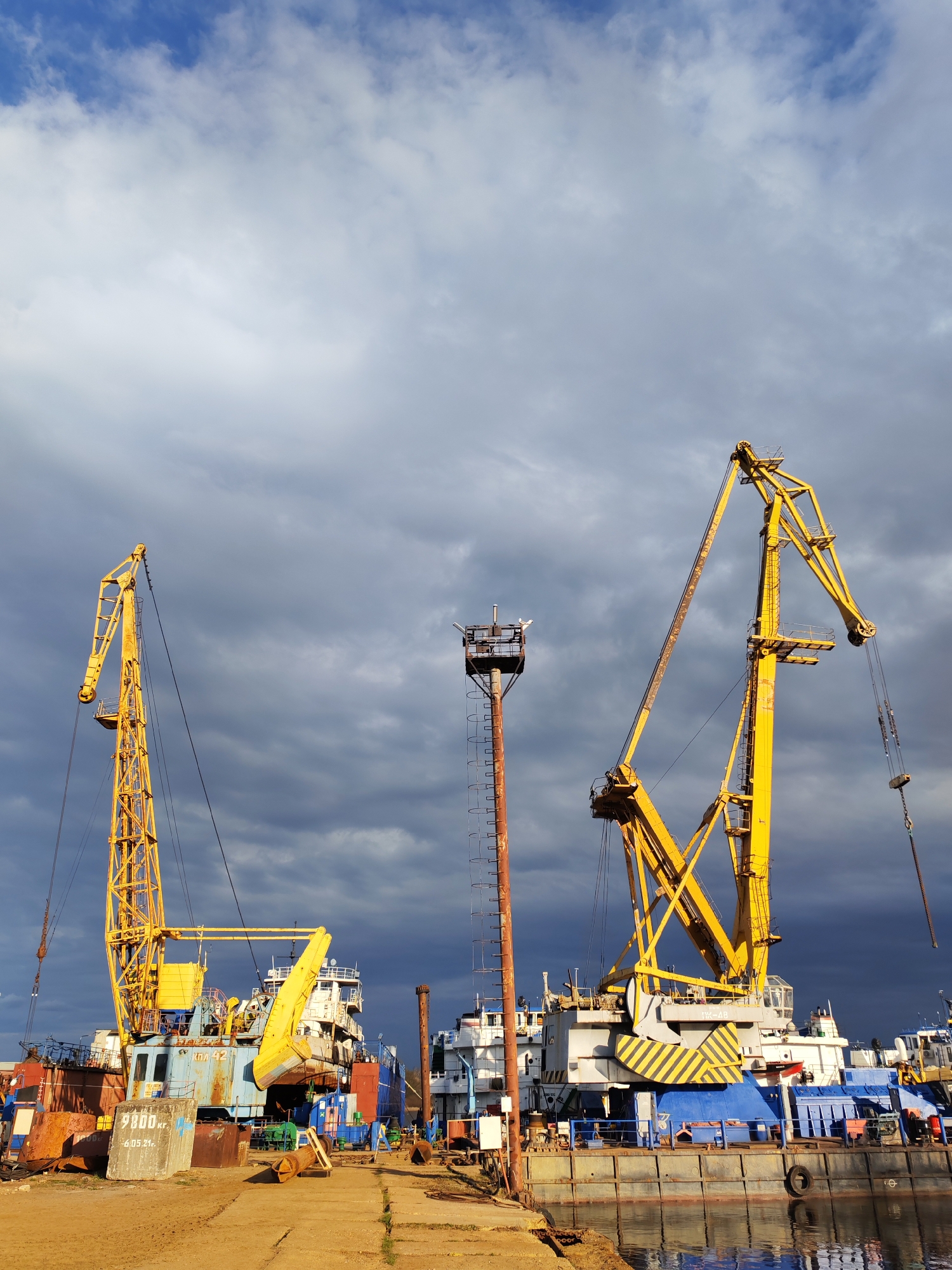 The image depicts a port scene in Rybinsk, Russia, with two large yellow cranes dominating the composition. The cranes are positioned on barges docked along the shoreline. A rusty metal tower stands between the cranes. The barges and surrounding area appear to be a working industrial site, with various pieces of equipment, vehicles, and materials visible. The sky is overcast with dark, dramatic clouds.