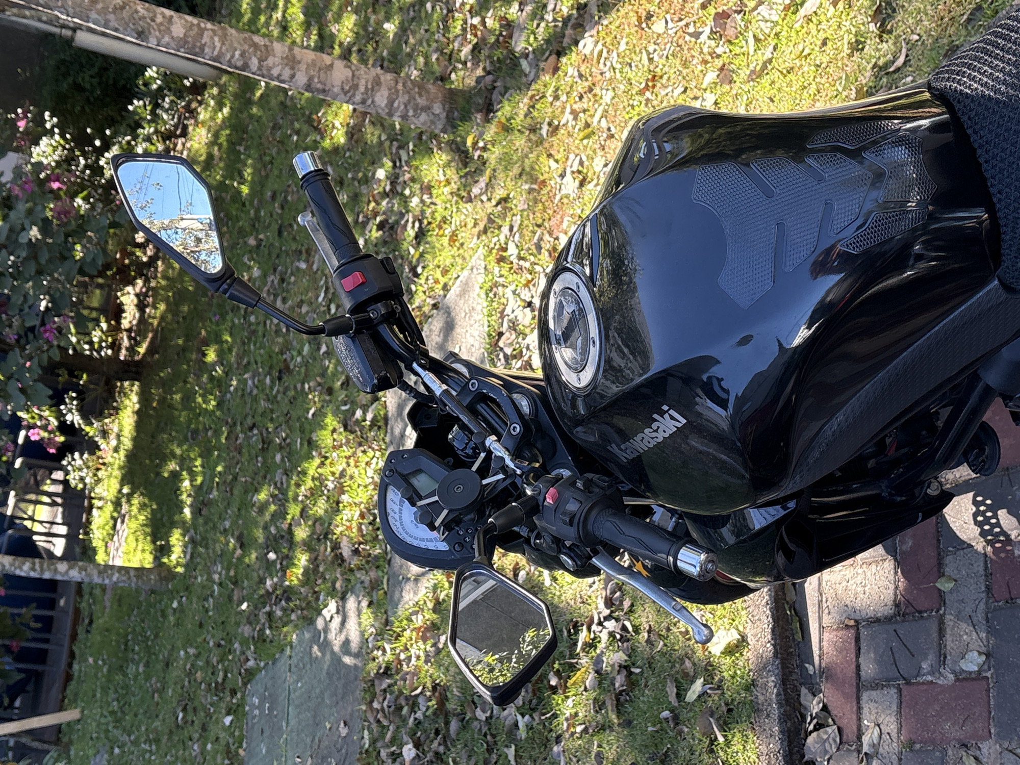 The image shows a black Kawasaki motorcycle parked outdoors on a sunny day. The motorcycle is positioned at an angle, with the handlebars and fuel tank prominently featured. The background consists of a grassy area with fallen leaves, trees, and some flowering bushes, suggesting a park or residential area. A paved walkway made of bricks is visible in the foreground.
The motorcycle is in good condition and appears to be parked, with no rider present. The mirrors reflect the sky and surrounding greenery. The fuel tank has a textured grip pad applied. The motorcycle's dashboard, with its speedometer and other indicators, is visible. The overall scene suggests a peaceful outdoor setting.