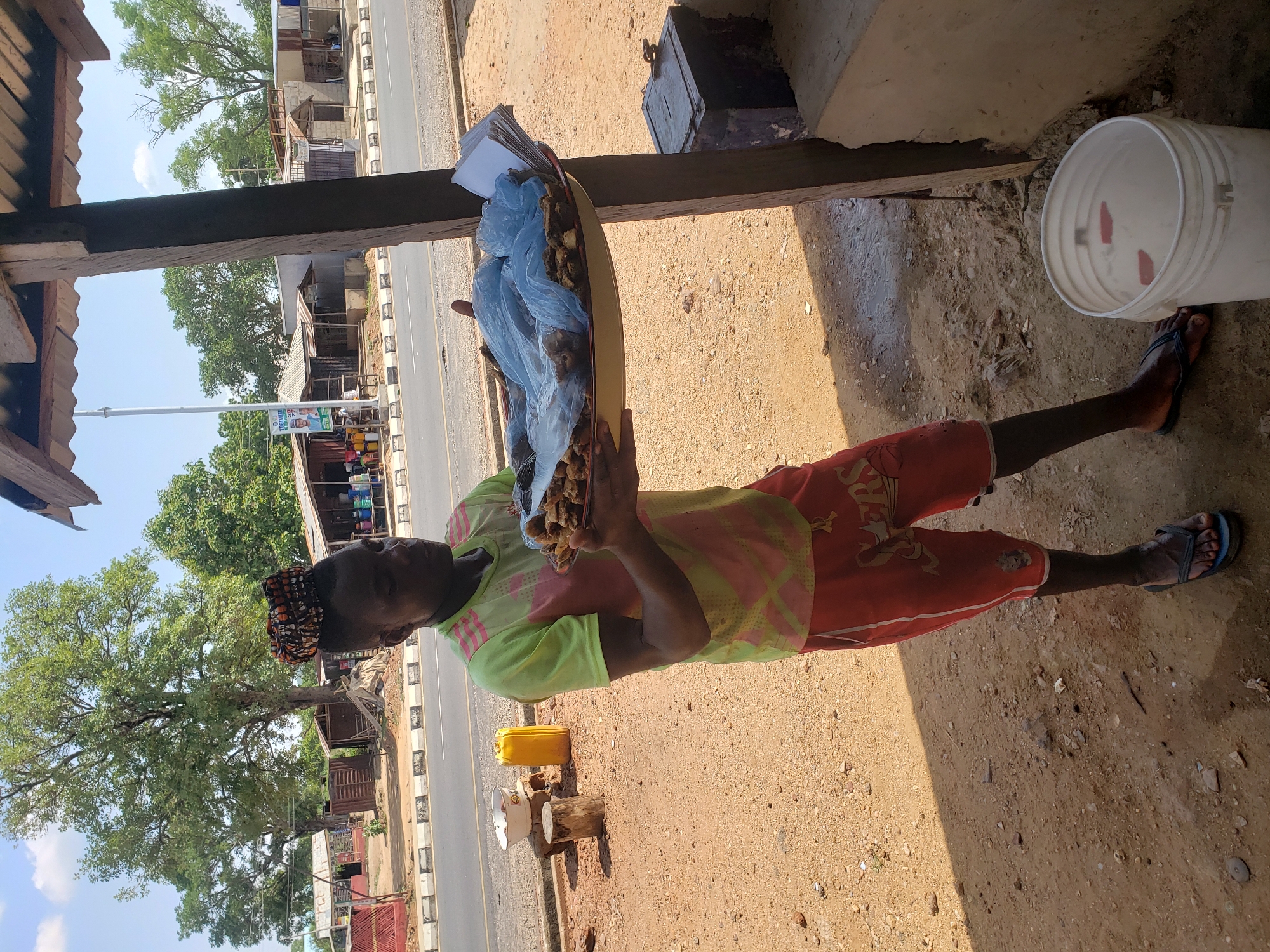 In Adankolo, Nigeria, a man stands beside a road under the shade of a building. He holds a platter filled with items packaged in blue plastic bags and some papers on top. He is wearing a light green shirt, red shorts, and sandals. A bucket sits next to him, and a yellow jerrycan can be seen nearby. The road is visible in the background with buildings and trees alongside it.