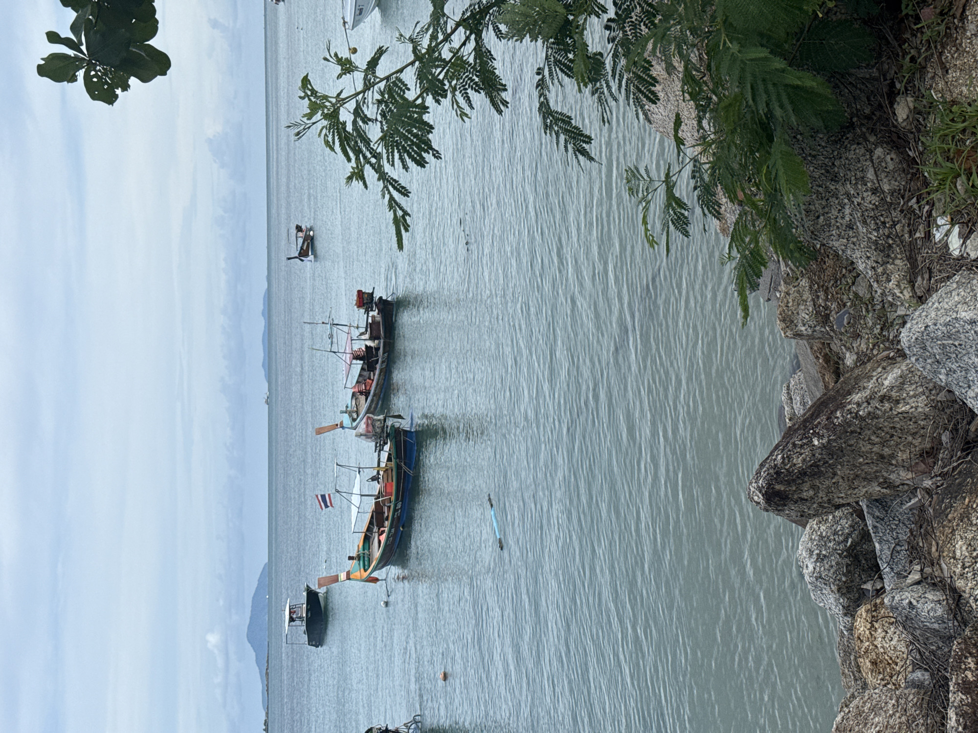 The image captures a serene coastal scene in Rawai, Thailand, under an overcast sky. The foreground is dominated by a rocky shoreline, with large, weathered stones of various textures and colors creating a rugged barrier. A few sparse green plants are growing amongst the rocks.
The middle ground features the calm, blue-green waters of the sea, with gentle ripples on the surface. Several boats are anchored or drifting in the water. Two prominent long-tail boats, characteristic of Thai fishing vessels, are positioned centrally. The boat on the left displays a Thai flag, and both boats have colorful wooden hulls and seating areas. Other smaller boats and a distant white vessel are visible further out at sea. A bright orange buoy floats near the shore, and a long, blue object, possibly a floating tube or pipe, is partially submerged in the water.
In the background, the horizon is marked by a hazy outline of islands or a distant coastline. The sky is a uniform shade of gray with scattered white clouds, suggesting a mild, possibly humid, climate. The overall atmosphere is one of tranquility, with the boats and the natural elements of water and rocks defining the picturesque location. No people are directly visible in the image.