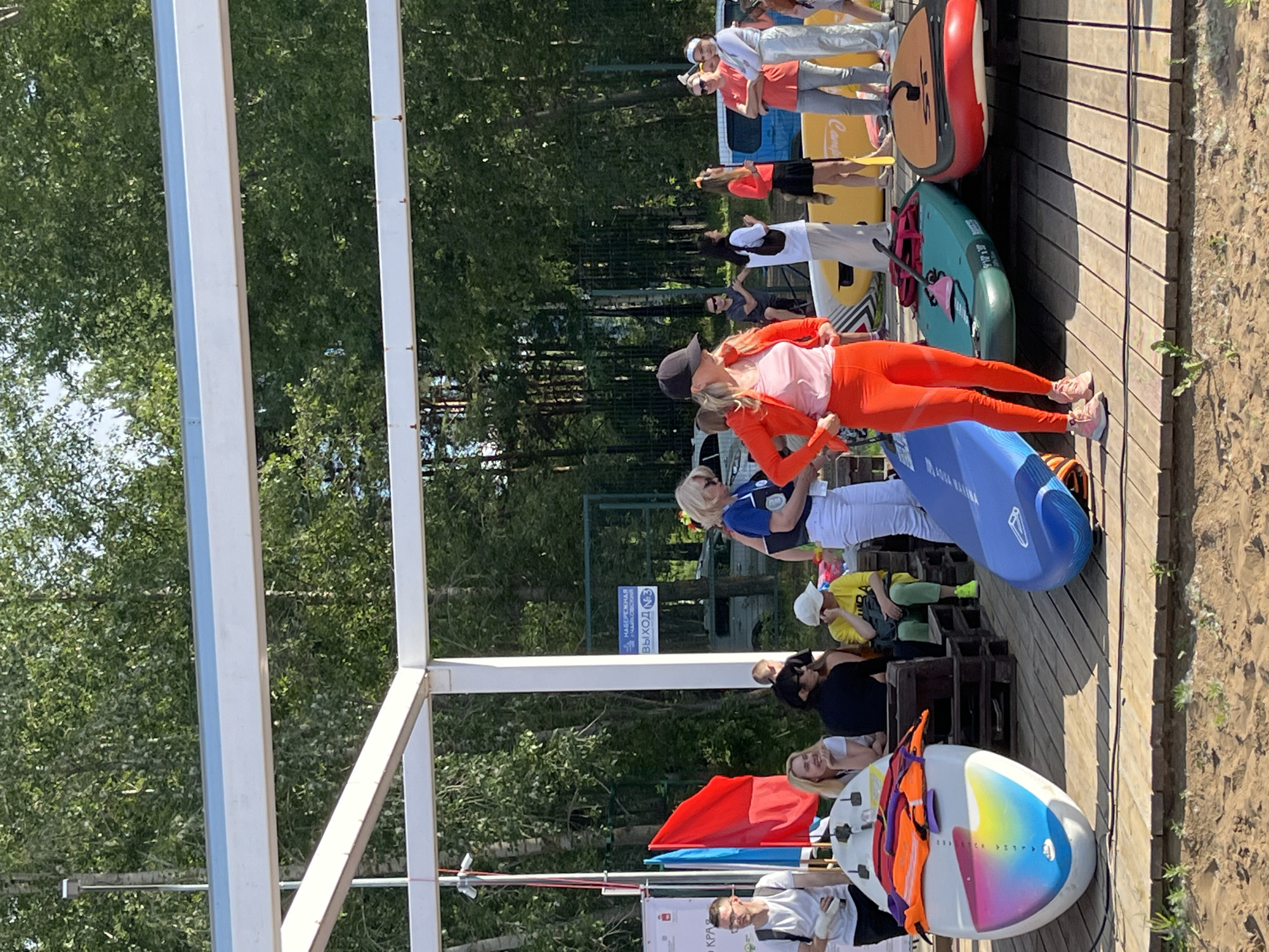 The image shows a group of people standing on a wooden deck near a body of water, likely a river or lake, surrounded by trees. The scene appears to be set outdoors during the daytime, with bright sunlight casting shadows. The weather seems pleasant, suitable for outdoor activities.
Several individuals are visible, some wearing casual clothing like t-shirts and pants, while others are in athletic wear. There's a woman in bright orange athletic leggings and top, and another in a pink shirt and orange jacket. A young boy in a yellow t-shirt and green pants is also present, sitting on some wooden pallets. A man in a white t-shirt and glasses is seen at the bottom of the frame.
Various objects, primarily stand-up paddleboards (SUPs), are arranged on the deck. One is a large, red and tan inflatable board. Another is a teal SUP with fins. A bright blue SUP is also prominent. A white SUP with colorful abstract art is at the bottom right, with an orange life vest draped over it. A red flag is also visible.
The people seem to be preparing for or participating in a paddleboarding event or activity. Their postures suggest a relaxed atmosphere, possibly a group gathering or lesson. The presence of multiple SUPs and life vests points towards water sports.
There is some visible text in the image. A blue sign with white lettering reads "НАБЕРЕЖНАЯ ВЫХОД N3". A white banner at the bottom has text in Russian, including "КРА". The overall setting appears to be a recreational area or a waterfront venue.