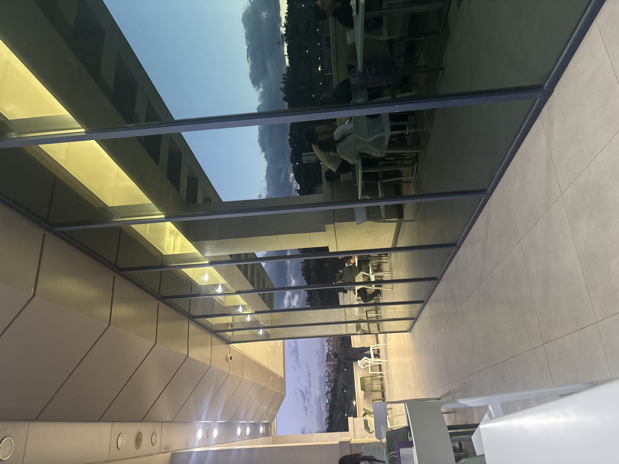 This image captures a modern architectural space, likely a covered terrace or a glass-enclosed lounge, at what appears to be dusk or early evening. The scene is dominated by extensive glass panels that form a ceiling and part of a wall, revealing both the exterior environment and reflections of the interior.
Through the glass, a soft, diffused sky with visible clouds stretches above a distant landscape of rolling hills and what seems to be urban development. Several individuals are visible through the glass, seated at tables with chairs, suggesting a dining or socializing activity. Their postures indicate relaxation, with one person standing near a table.
The interior architecture on the left features distinctive light-colored, angled panels with circular embedded lights, casting a warm, yellowish glow within the space. The floor, visible in the lower right, is composed of large, light-colored square tiles. White and some green chairs, along with various tables, are arranged both within the main viewing area and beyond the glass. The reflections in the glass show elements of the building's structure and interior lighting. No text is visible in the image. The setting suggests a contemporary venue in Yerevan, Armenia.