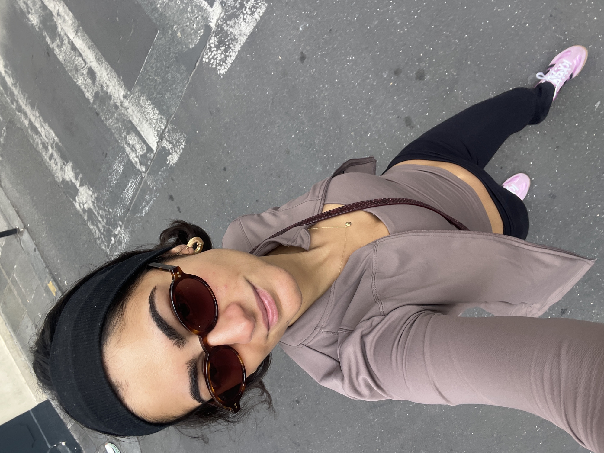 A woman is standing on a city street, possibly in Paris, France, as indicated by the markings on the asphalt. She is wearing a black headband, round sunglasses, a mauve-colored athletic jacket, a matching top, and black leggings. She has pink shoes with white laces on her feet. The surface beneath her feet is gray asphalt with visible road markings. She is looking directly into the camera with a slight smile.