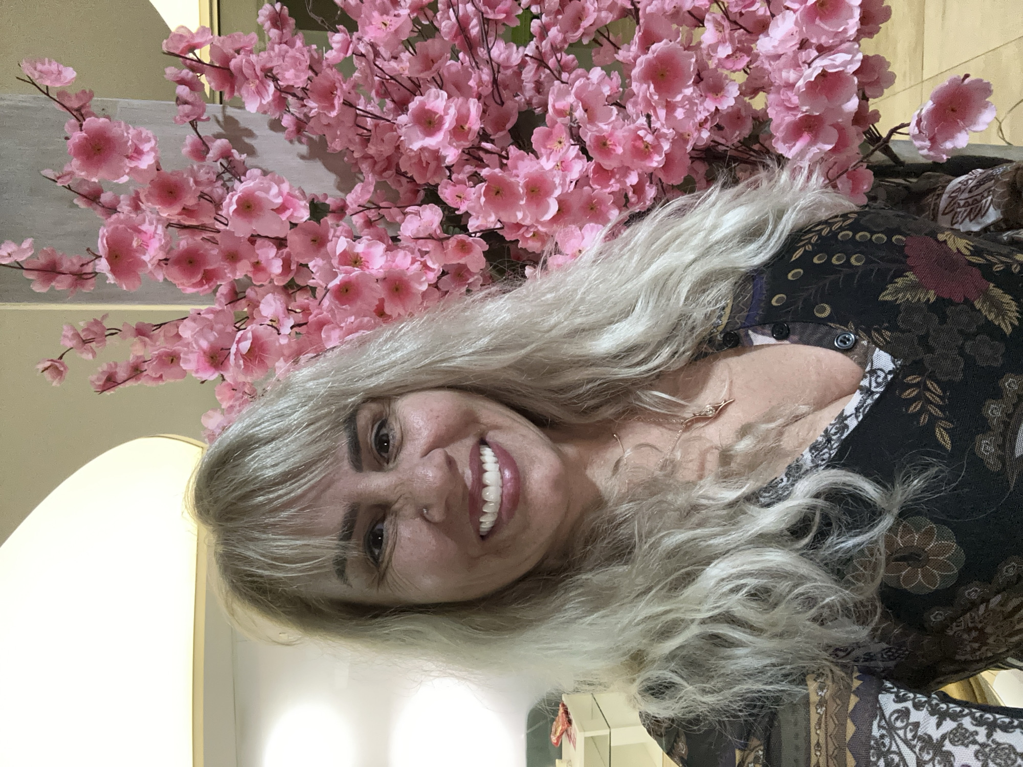 A woman with long, wavy, light blonde hair is captured in a cheerful, upright portrait, smiling broadly towards the camera. Her dark eyes crinkle at the corners, and her wide smile reveals white teeth. She has bangs across her forehead. She wears a dark top or dress adorned with a complex pattern of abstract and floral designs in hues of black, brown, and deep red. A delicate necklace with a small pendant is visible around her neck.
Behind and to her right, a vibrant cluster of artificial pink cherry blossoms dominates the background, with numerous branches and individual flowers creating a lush, decorative effect. Parts of a light-colored, possibly off-white, interior wall or structure are visible behind the blossoms and to the woman's left, indicating an indoor setting. A bright, undefined light source or reflective surface is present in the lower left portion of the frame.
The image conveys a sense of happiness and pleasantness, with the woman appearing to pose for the photograph in a decorated indoor environment, possibly in Sofia, Bulgaria, as per the location context. There is no visible text, and the time of day is not discernible from the indoor lighting.