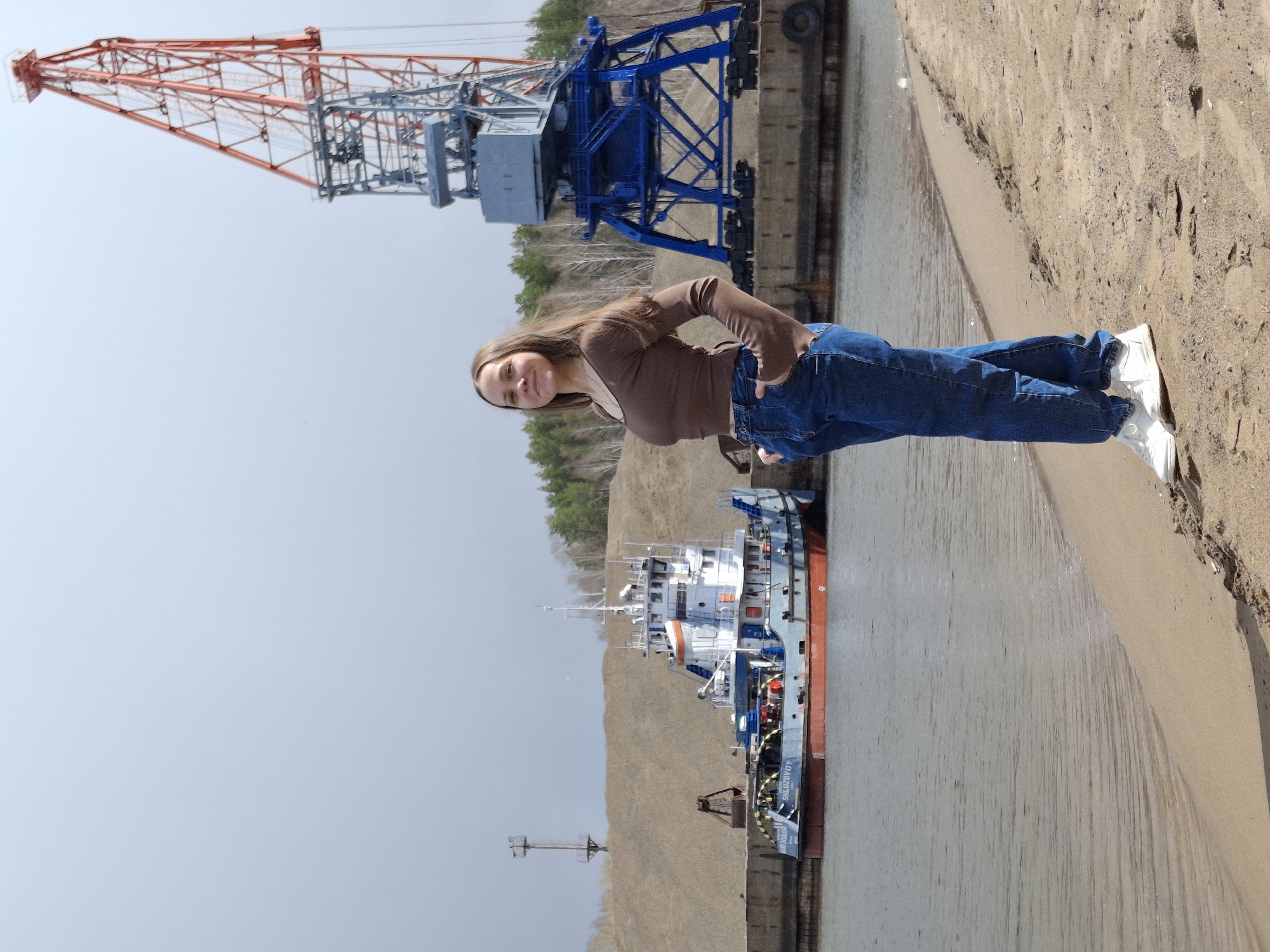 This image, set in Chaikovsky, Russia, depicts a vibrant industrial waterfront scene on a clear, sunny day. The main subject is a young woman, likely in her late teens or early twenties, with long brown hair, who stands smiling at the camera. She is dressed casually in a brown long-sleeved top, blue jeans, and white sneakers, with her hands in her pockets, suggesting a relaxed pose.
Behind her, the scene unfolds with industrial elements. A large, prominent gantry crane, painted red and blue, dominates the upper left portion of the background. Below this, a blue industrial crane or structure is fixed on a dock or embankment. To the right of the woman, a body of water, likely a river, stretches out, leading to a sandy or dirt bank on the far right.
Further in the background, a white, blue, and red boat or small ship is moored or docked. The boat has visible text on its hull: "БАЛТИЯ" (BALTIYA) and "ОАО ДЗТНЗ". Beyond the boat and the water, a distant landmass with sparse trees and what appears to be a low, brownish-green hill or embankment is visible under a bright, light blue sky. The overall mood is casual and bright, with the woman appearing cheerful against the busy backdrop of a working port.