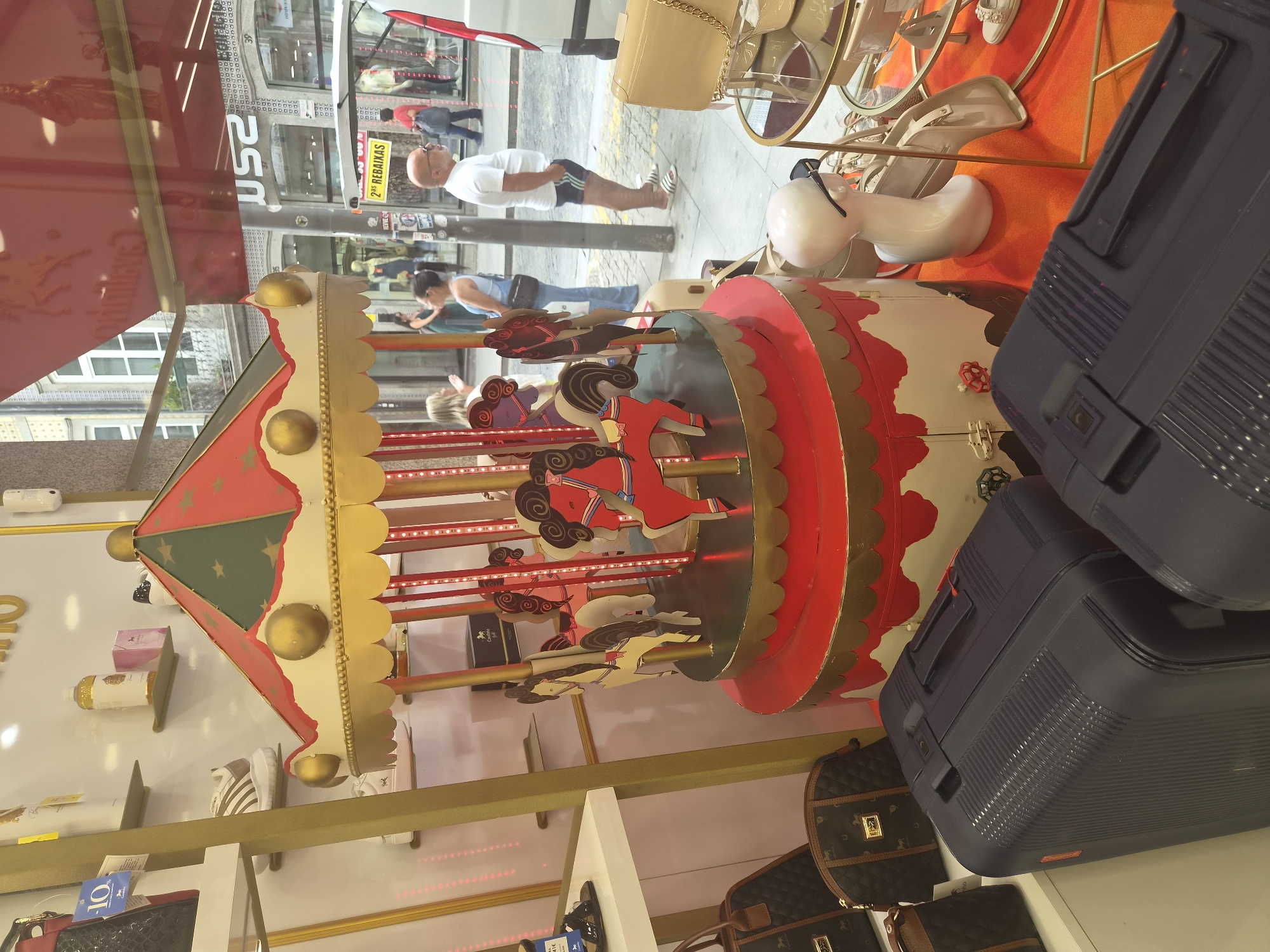 In Braga, Portugal, a store window displays a festive carousel with red and gold detailing and carousel horses. In the foreground, two dark blue suitcases are visible. Behind the carousel, through the glass, people are seen walking on a street with shops. A yellow sign indicating "2a REBAIXAS" (2nd Discounts) is attached to a pole outside. A white van is parked to the right. The scene captures a retail environment with seasonal decorations and a glimpse of the urban surroundings.
