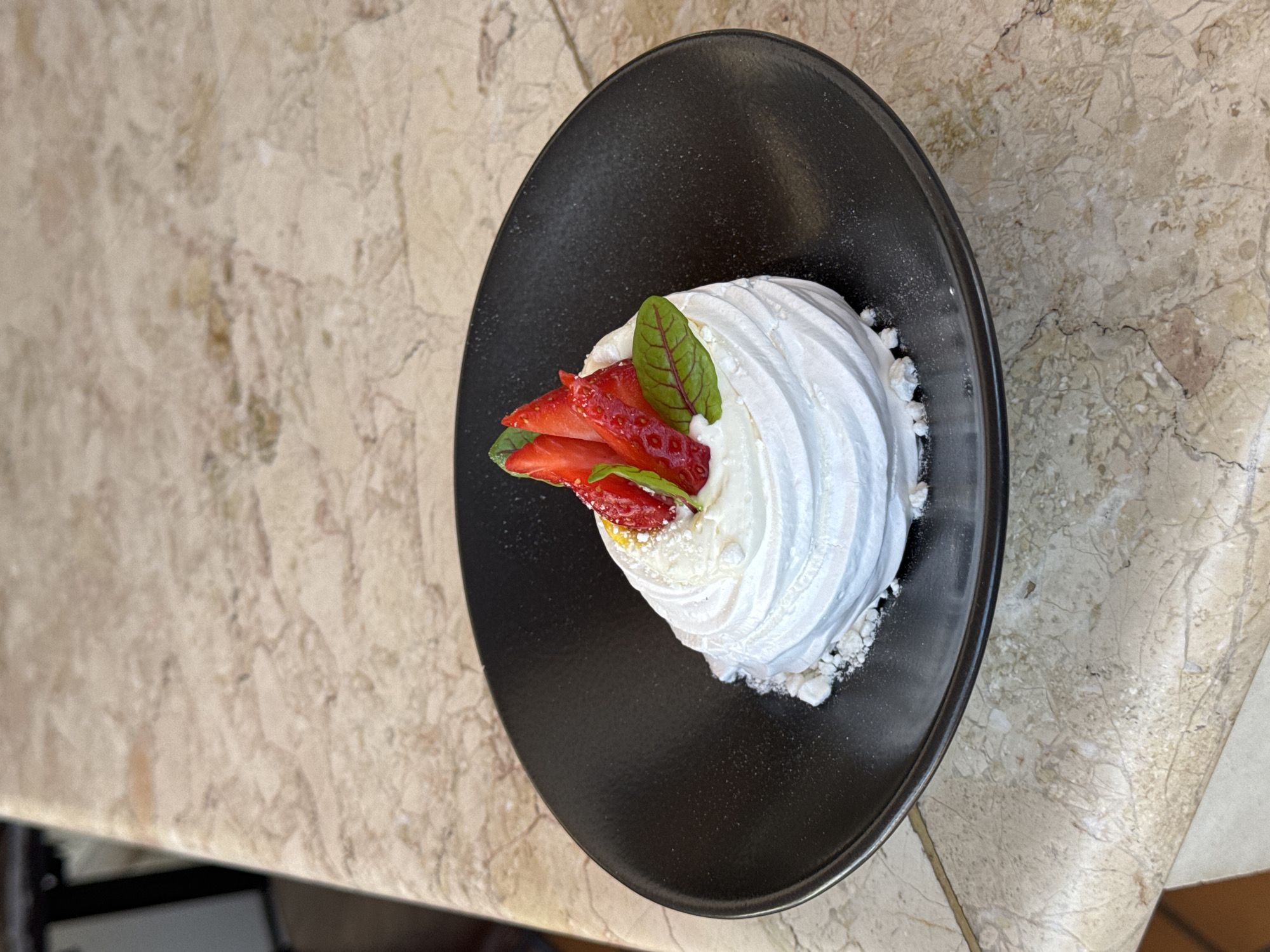The image shows a dessert, likely a pavlova, served on a matte black plate. The pavlova is topped with sliced strawberries and two small green leaves. Crumbs of meringue surround the base of the pavlova on the plate. The plate rests on a light-colored, faux-marble surface. The location appears to be in Sochi, Russia. No people are visible.