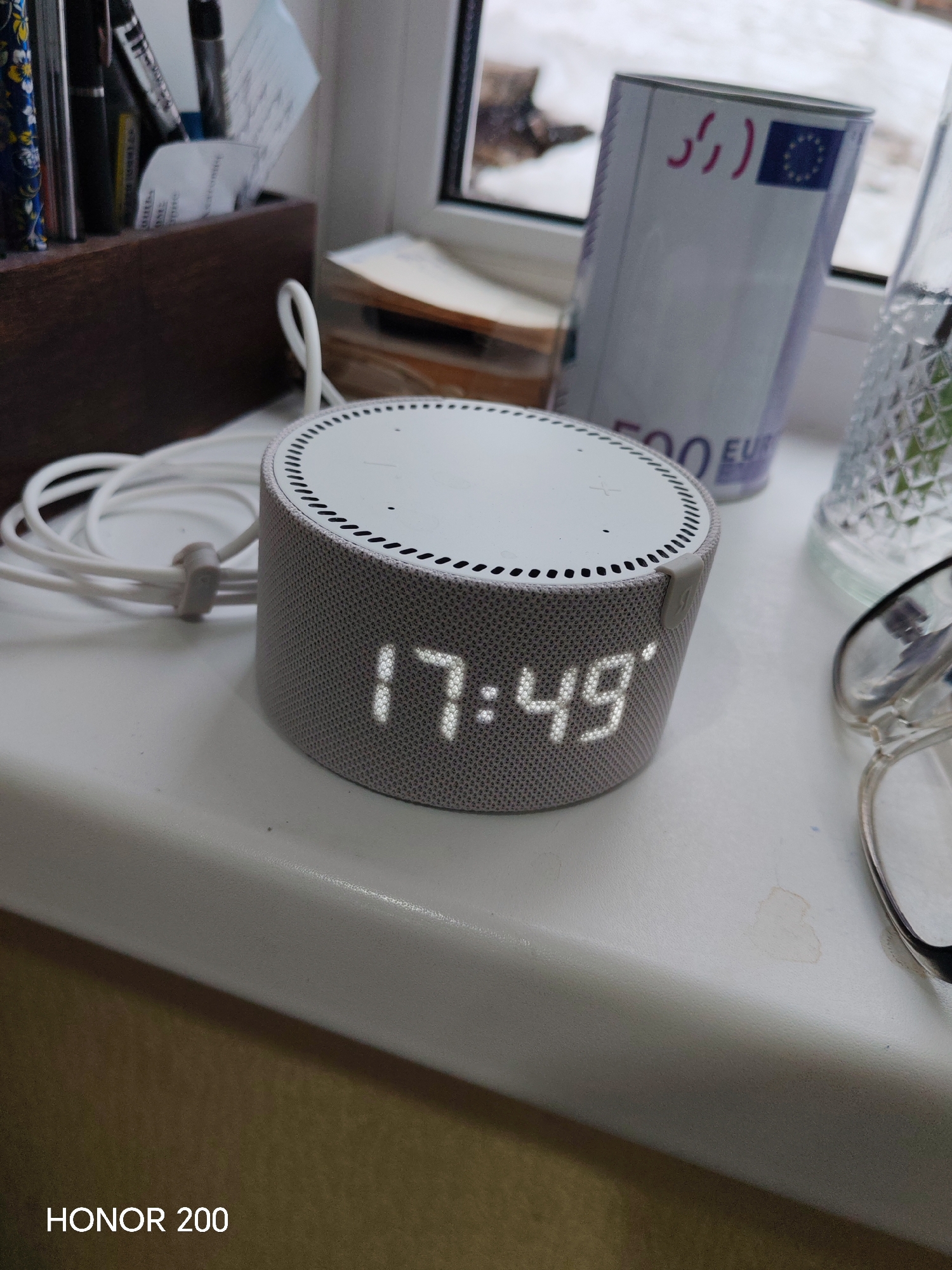 On a white windowsill, a gray smart speaker displays the time 17:49. To the left of the speaker, a white charging cable is coiled. Behind the speaker are a wooden pen holder filled with pens and pencils, a stack of books, a novelty 500 Euro bill-themed container, and a glass with a geometric pattern. To the right of the speaker are eyeglasses. Outside the window, snow is visible. The lower-left corner of the image includes the text "HONOR 200". There is no indication of the location being Izhevsk, Russia.