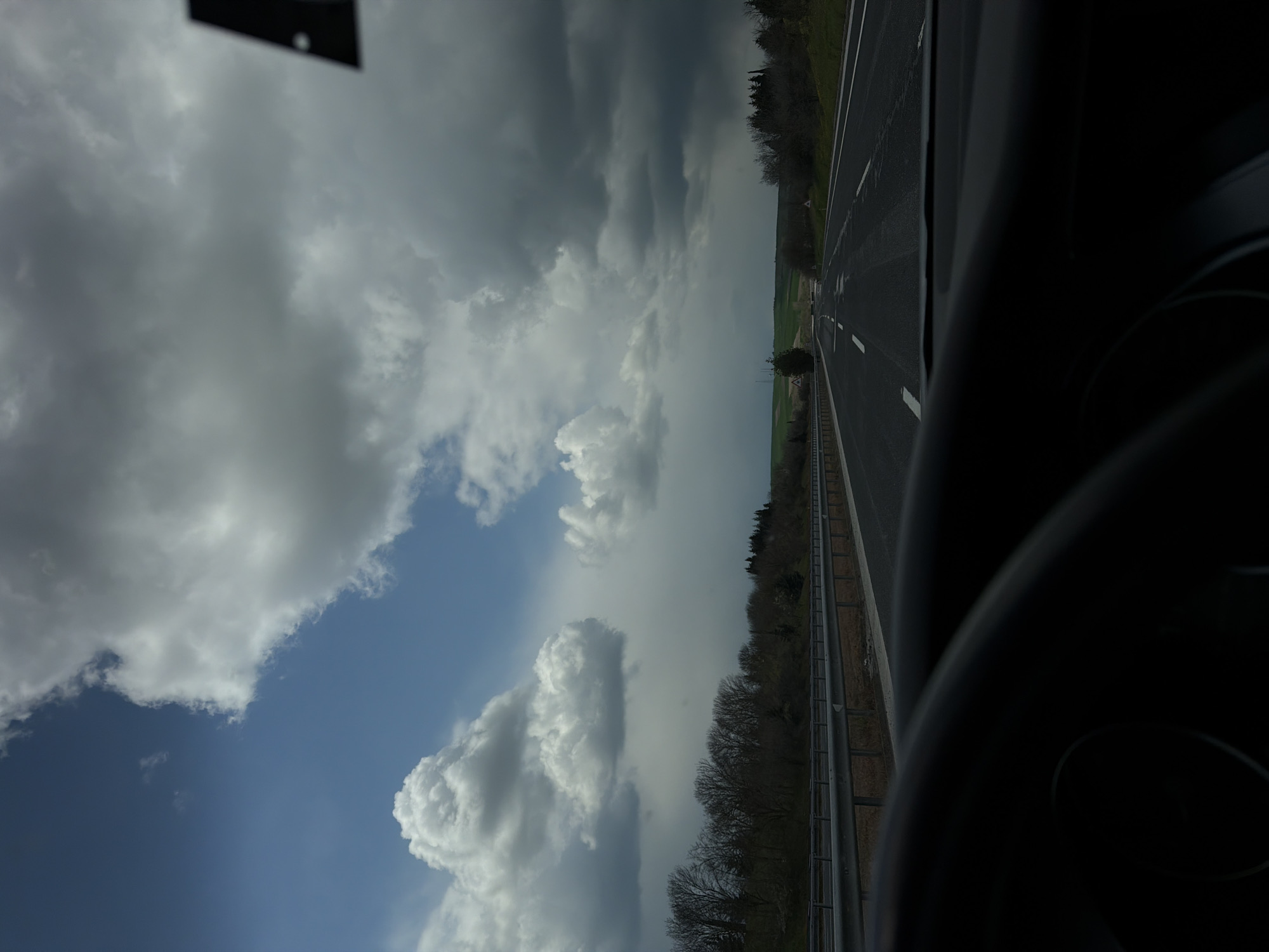 The image shows a view from inside a vehicle, likely a car, looking out onto a highway. A portion of the steering wheel is visible on the right side of the frame. The highway stretches into the distance with visible lane markings. A line of trees and a grassy area border the road. The sky is filled with clouds, with patches of blue visible. The overall lighting is subdued, suggesting an overcast day.

While the image depicts a highway scene, it is not possible to confirm its location specifically in Havsa, Turkey without further information or context.