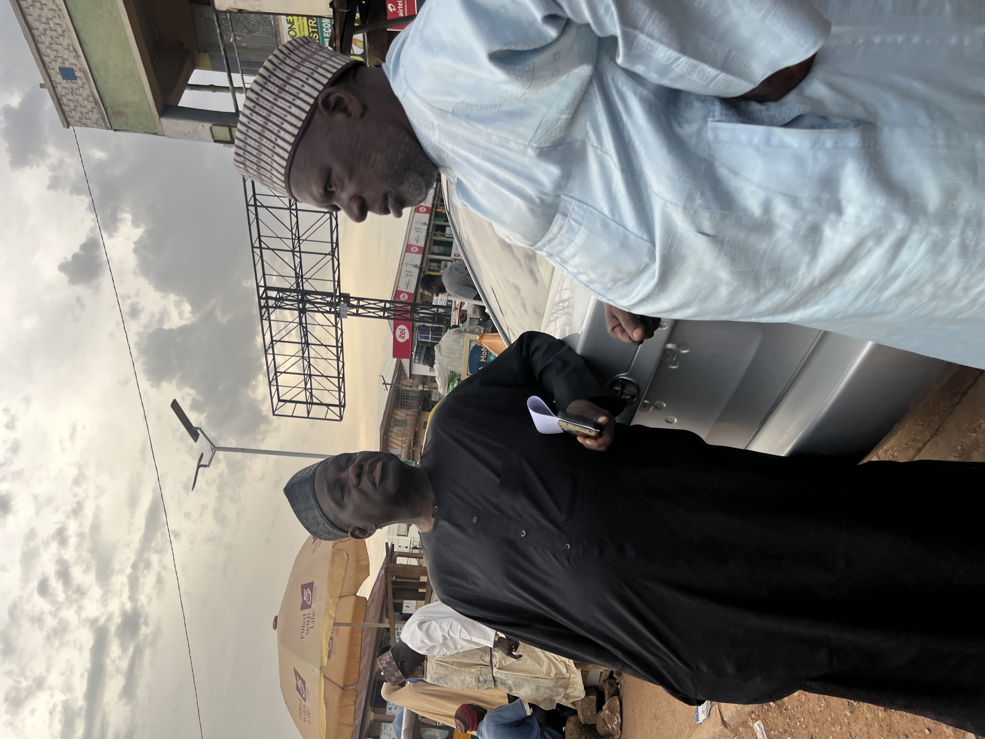 The image depicts two men engaged in conversation next to a silver car on a street in Bukuru, Nigeria. The man on the left, wearing a black traditional outfit and a dark cap, is looking towards the right. He is holding a folded paper in his right hand. The man on the right, dressed in a light blue traditional outfit and a striped cap, is facing the man in black and appears to be speaking.
The environment suggests an outdoor setting with elements of a marketplace or street vendor area. An yellow umbrella with a logo is visible behind the man on the left, along with stalls and buildings in the background. The sky is overcast with clouds. Other individuals and vehicles are partially visible in the background, indicating a public space with ongoing activity. The ground is dirt and gravel, typical of many Nigerian roads. The overall impression is that of a casual encounter or discussion taking place in a bustling urban environment.