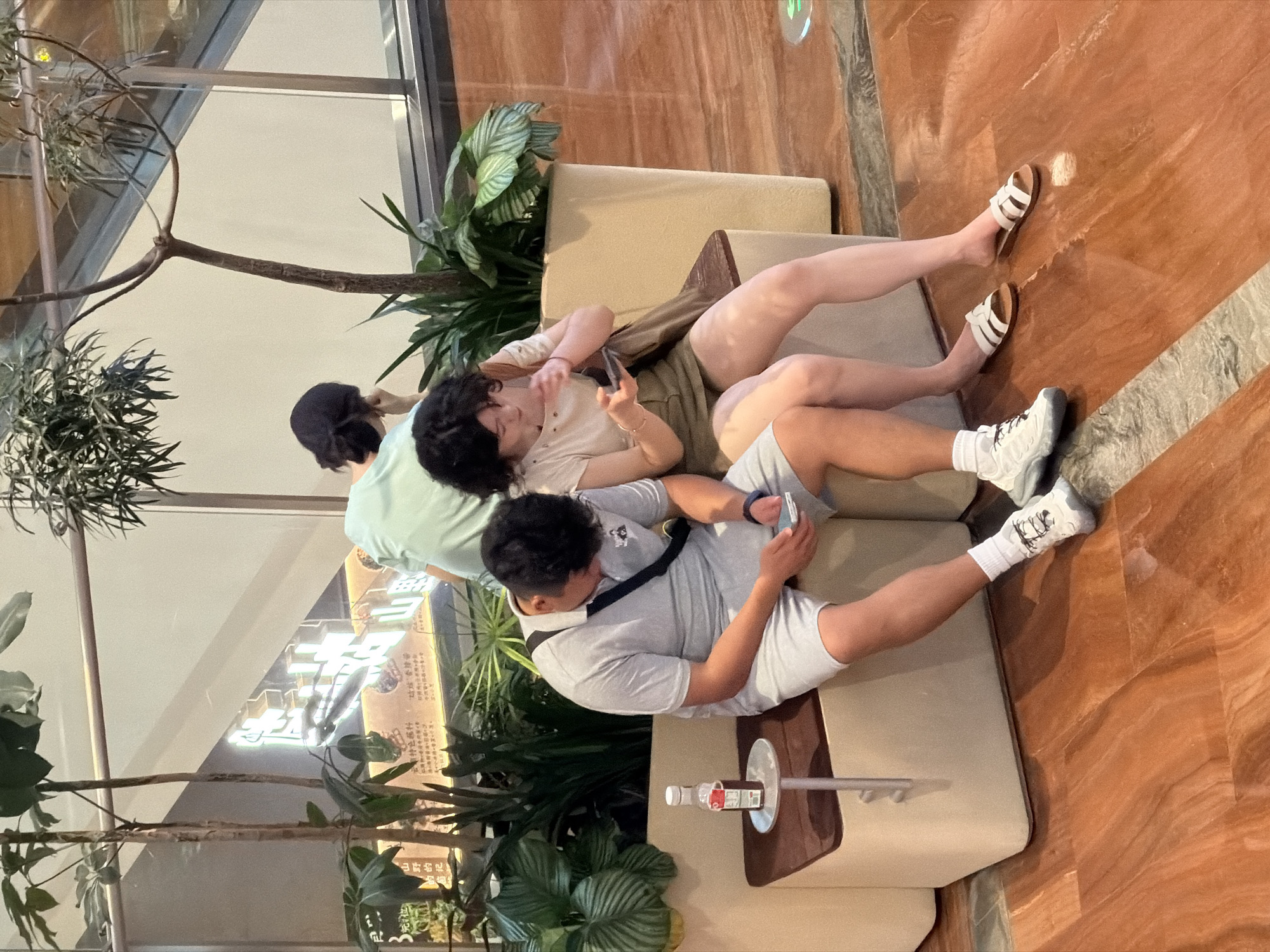 The image shows three individuals sitting on a light-colored bench in an indoor environment with polished wood-like flooring. In the foreground, a man wearing a light blue polo shirt and shorts is looking down at his mobile phone. Beside him, on his left, is a small side table with a bottle of dark liquid. To his right, a woman wearing a light-colored t-shirt and olive-green shorts is also looking at her mobile phone, with her arm resting on the man's shoulder. Behind them, a third person wearing a light green t-shirt and a dark cap is partially visible, leaning forward. The background includes potted plants and a staircase with illuminated signage that appears to be in Chinese characters, suggesting the location is in Yuzhong District, China. The overall lighting suggests an indoor commercial or public space.