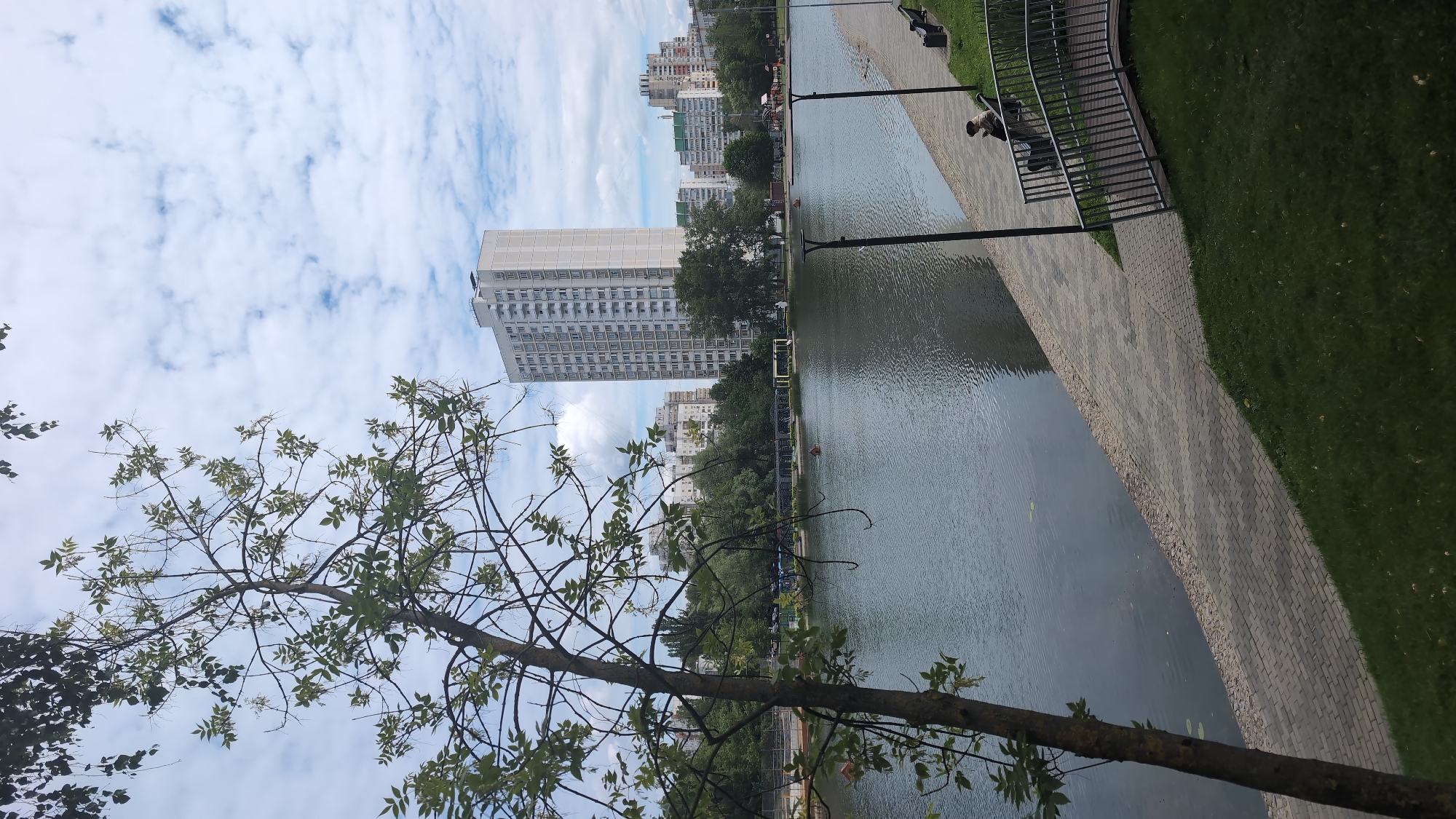An eye-level, medium shot captures a serene view of a pond in Moscow, Russia, on a cloudy day. The foreground is dominated by the lush green grass and the textured surface of a paved walkway that lines the pond's edge. A thin tree with sparse green leaves stands to the left, its branches reaching towards the sky. Beyond the walkway, the calm water of the pond reflects the overcast sky, with a few small objects floating on its surface.
Across the pond, a cluster of multi-story buildings forms the backdrop. The most prominent is a tall, light-colored residential building with numerous windows, standing out against the other, slightly shorter buildings. A line of trees separates the pond from these structures, adding a touch of nature to the urban landscape. The sky is a canvas of white, fluffy clouds scattered across a pale blue expanse.
Further along the walkway, two dark-colored benches are visible, with one occupied by a person wearing a dark jacket and a hat, seemingly lost in thought or enjoying the view. A sleek metal railing forms a bridge-like structure, adding a modern architectural element to the scene. The overall impression is one of quiet contemplation amidst a bustling city.