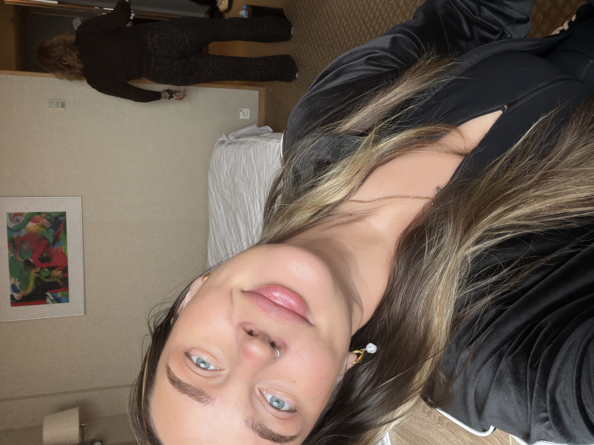 The image is a selfie of a young woman with fair skin and light blue eyes, taken in a room. She has long, light brown hair with blonde highlights. She is wearing a black robe or jacket with white piping around the edges. She has a nose ring and an earring with a pearl on it.
In the background, there is another person wearing a black outfit with an animal print on the pants, bending over or leaning against a wall. The wall is a neutral beige color. There is a framed piece of art hanging on the wall, and a lamp can be seen near the bottom left corner of the image. A white bed with textured bedding is also visible.
It is not possible to determine whether the location is in the city of Moscow, Russia, or any other specific location, based on the image.