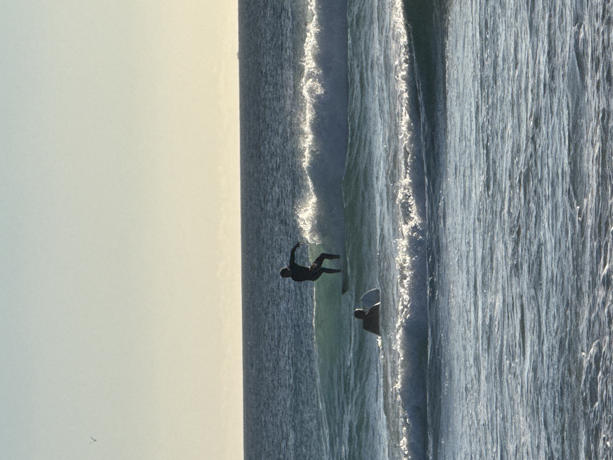 The image shows two surfers riding a wave in Hout Bay, South Africa. The sky is light-colored, possibly indicating sunrise or sunset. The water is choppy with whitecaps. One surfer is further up the wave and slightly airborne, while the other is positioned further down and behind. The image is rotated.