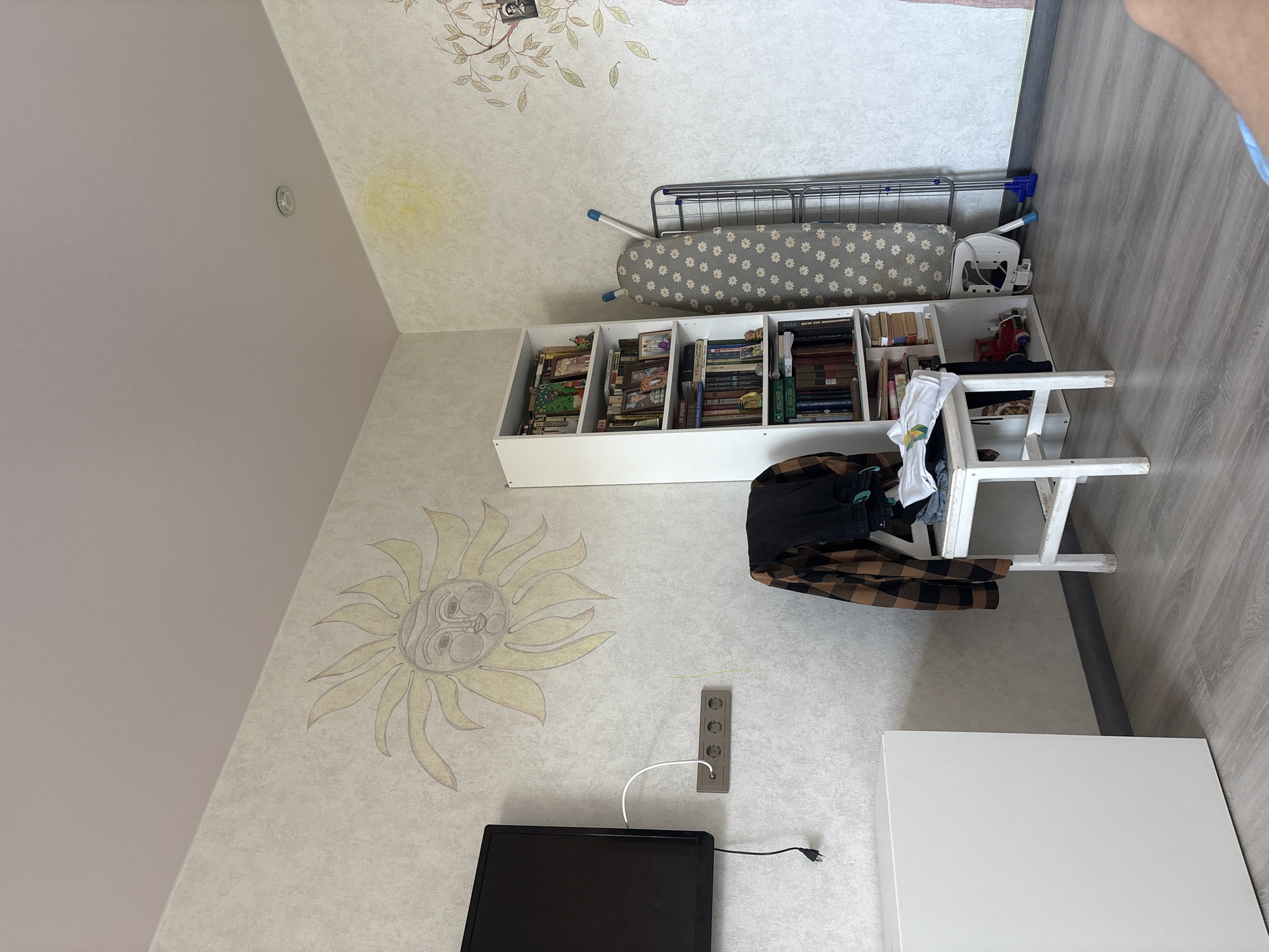 The image shows an indoor scene, likely a bedroom or living area. The walls are textured and light-colored, with a sun mural on the left and a partial tree mural on the right. A flat-screen television is mounted on the left wall, connected to a white cable that runs to an electrical outlet strip below. To the right of the television, a white cube-shaped cabinet is visible. In the center of the room, a white wooden chair is placed in front of a white bookshelf. Clothing, including a plaid shirt and dark pants, is draped over the chair. The bookshelf is filled with books and decorative items, including picture frames. Next to the bookshelf, a gray ironing board with a daisy pattern is set up, with a metal drying rack beside it. The floor is covered with light gray laminate flooring. The ceiling is light pink and a smoke detector is visible in the upper right corner.