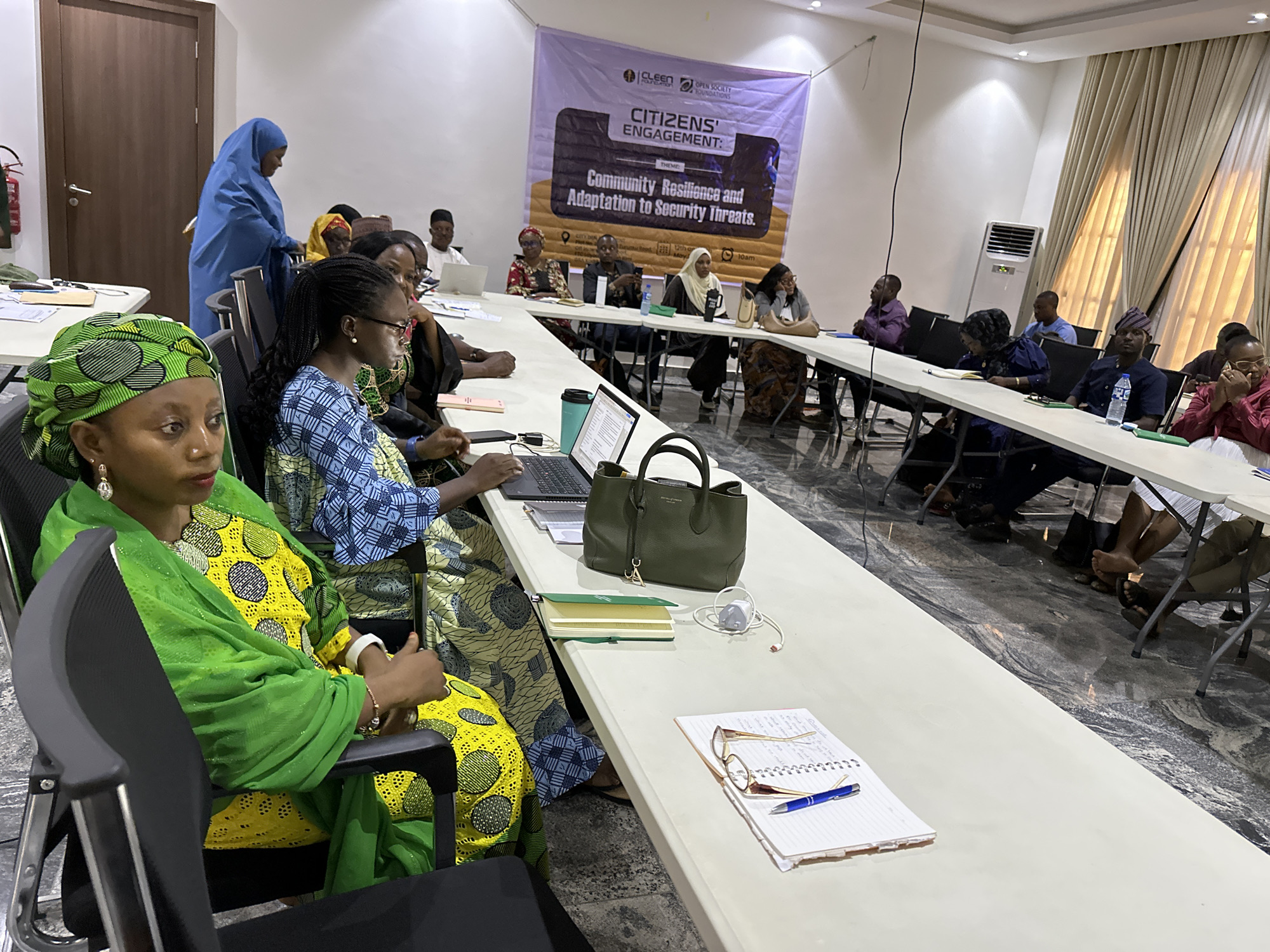 The image shows a conference room during a citizens' engagement event, likely in Unknown, Unknown. About 20 people are seated around a long, white table, participating in the event. A banner on the wall reads "Citizens' Engagement: Community Resilience and Adaptation to Security Threats," indicating the event's theme. The banner also features logos of the CLEEN Foundation and Open Society Foundations. Participants are a diverse group, mostly adults, seated along the table, some with laptops, notebooks, and other personal items in front of them. One woman in the foreground is wearing a green headscarf and a yellow patterned dress with a green shawl. Another woman, in blue, is standing to the side. The room has white walls, curtains, and an air conditioner in the background. The flooring appears to be patterned marble.