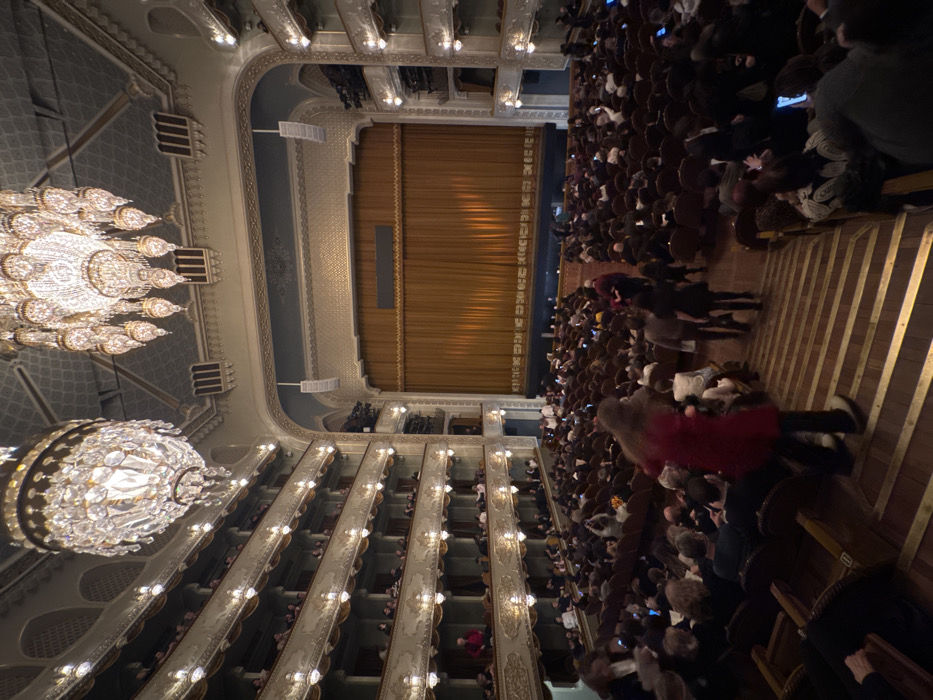 The image shows the interior of an ornate theater, viewed from a high vantage point. A large, crystal chandelier hangs prominently on the left. The theater is packed with an audience facing a closed stage curtain. Multiple levels of box seating are visible on both sides, filled with spectators. The architecture features intricate details and illuminated by numerous small lights.