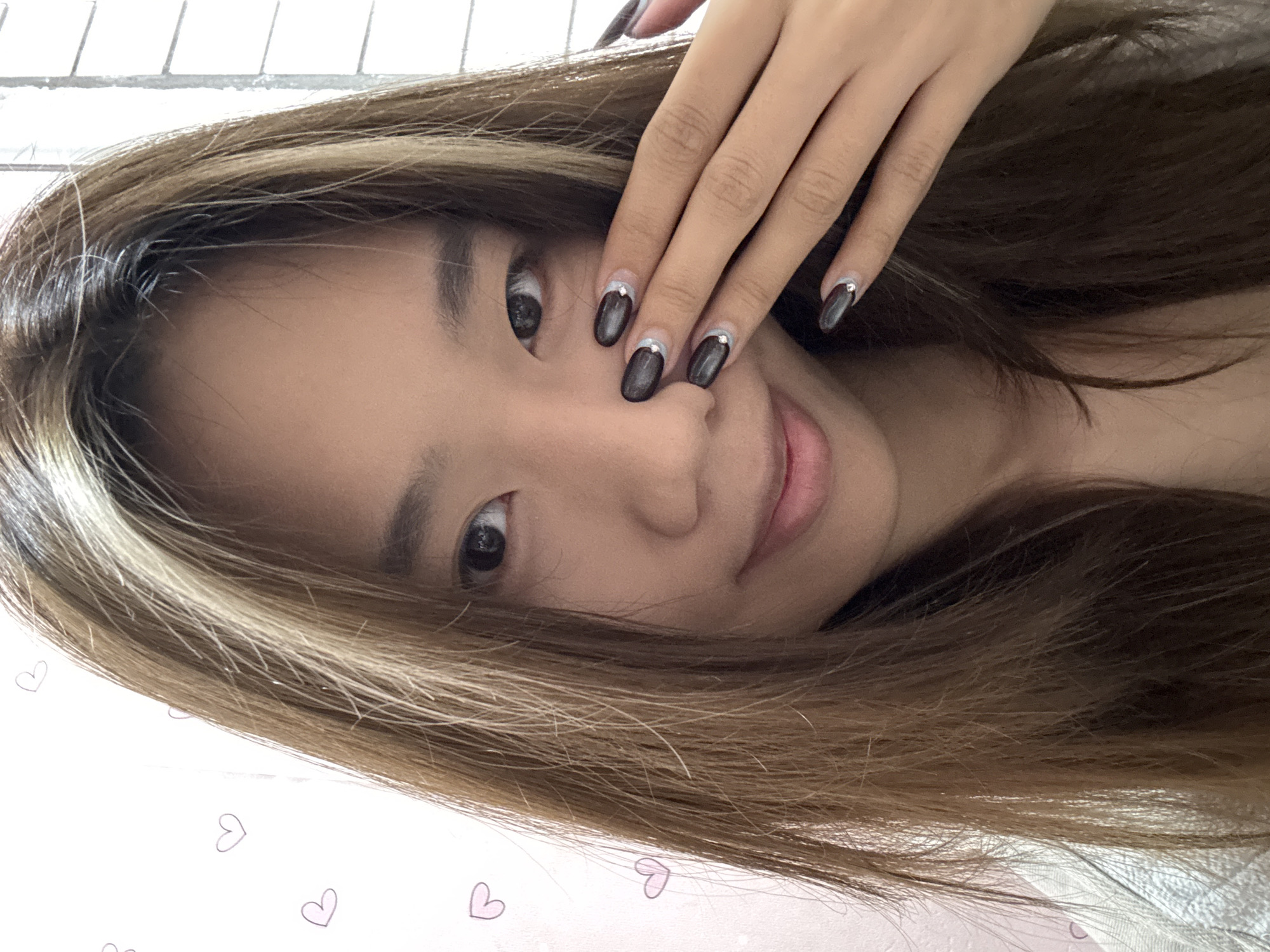 A young woman with tan skin and dark eyes is posed in a selfie-style image. Her light brown hair with blonde highlights frames her face, partially obscuring her left eye. She is smiling slightly. Her right hand is near her face, displaying dark-polished nails with silver accents near the cuticles. The background appears to be a white surface with faint pink heart patterns. There is no visual information in the image to confirm or deny the location in Shapingba District, China.