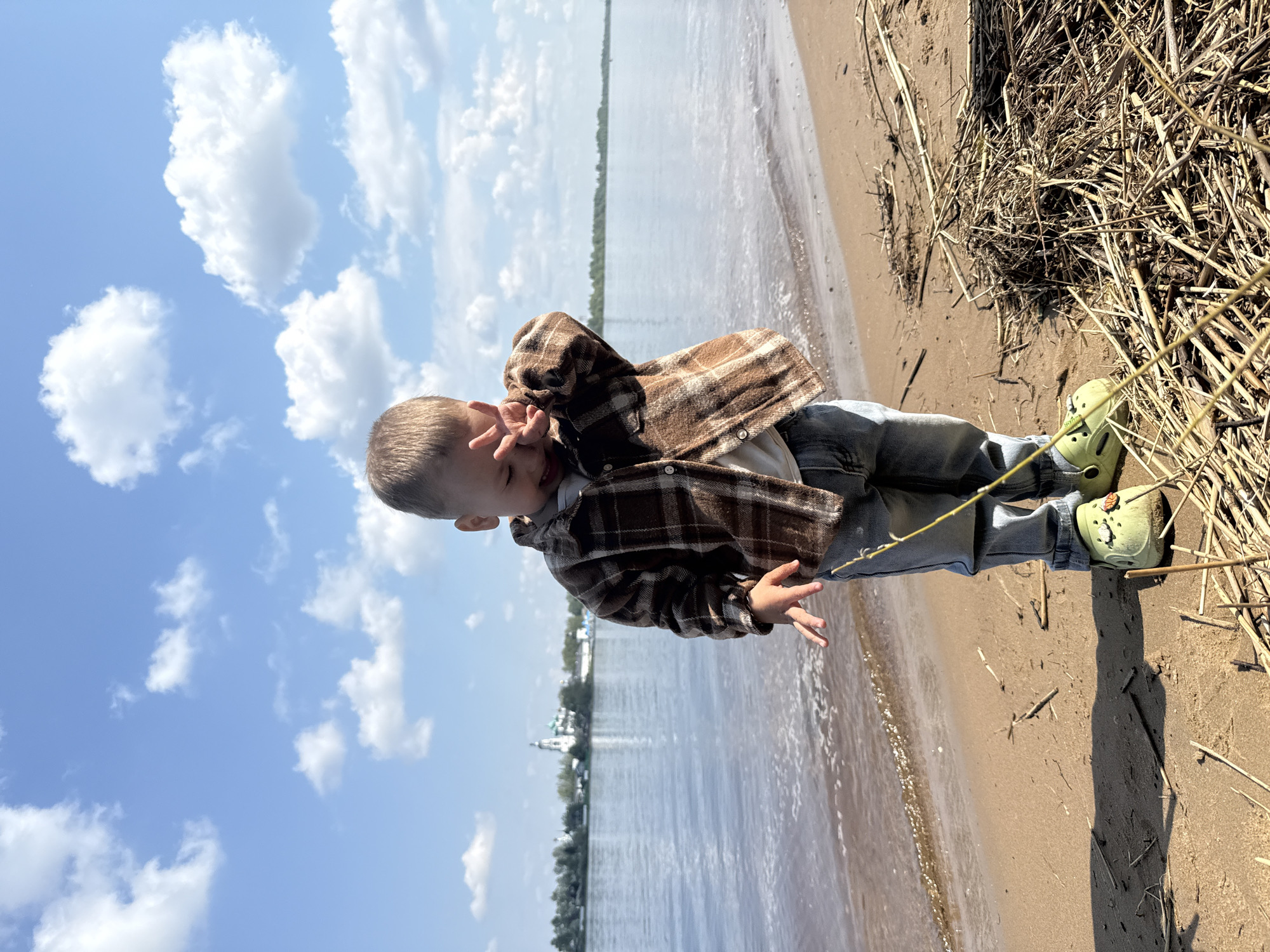A young child stands on a sandy beach near a body of water. The child is wearing a brown and tan plaid shirt, blue jeans, and green Crocs. The water has small waves lapping at the shore. The background shows a clear sky with some clouds, with buildings in the distance. The location is Rusanovo, Russia.