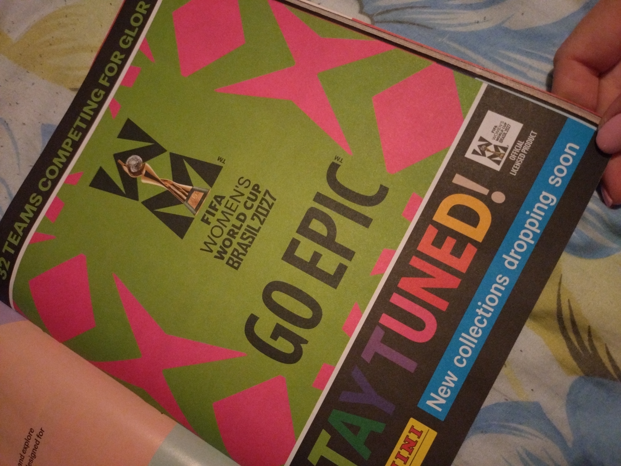 The image shows a close-up of a page from a magazine or brochure, with a prominent green background and large pink star shapes. Text overlays the image. At the top, partially visible, is the text "32 TEAMS COMPETING FOR GLOR". Below that, in the center, is a graphic of a trophy with the text "FIFA WOMEN'S WORLD CUP BRASIL 2027". Larger text reads "GO EPIC". In the lower half of the image, large, colorful letters spell out "STAY TUNED!". Below this, in a blue bar, is the text "New collections dropping soon". In the bottom left corner, a yellow rectangle contains the word "NINI". In the bottom right, a white square contains the FIFA Women's World Cup logo and "OFFICIAL LICENSED PRODUCT". The image is taken from an angle, with a hand visible in the bottom right corner, suggesting it's being held. The background texture appears to be fabric with a floral pattern. There is no visible indication of the city of Stara Zagora, Bulgaria.
