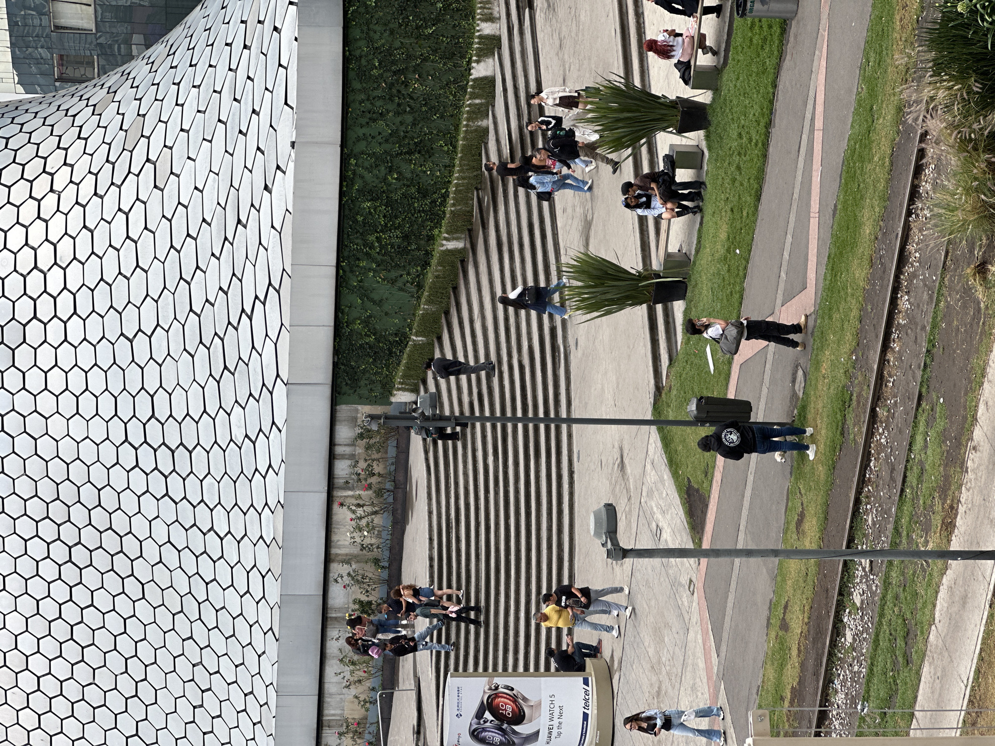 The image shows an aerial view of a public space in Mexico City, Mexico. The Soumaya Museum, a modern building with a unique hexagonal patterned facade, is visible on the left. Adjacent to the museum is a series of wide concrete steps interspersed with narrow garden beds containing low green plants and small flowering trees. People are walking on the steps and the pavement, including groups of friends, couples, and individuals. Some are sitting on benches. The area is separated from a road by a strip of grass. There are also lampposts and a billboard advertising a Huawei watch.