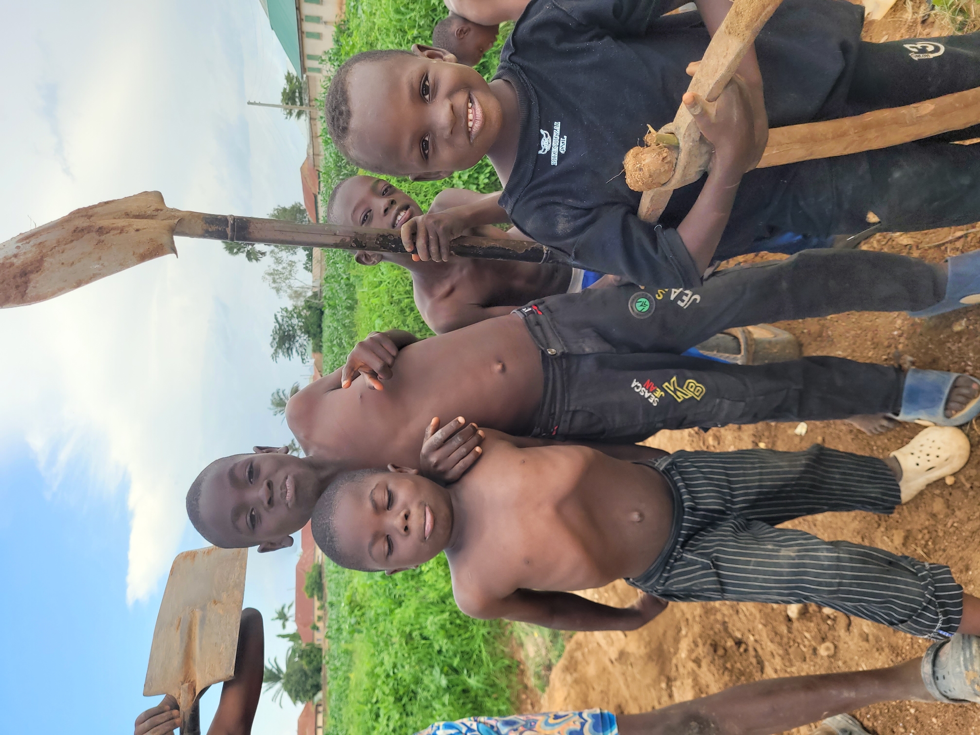 In Bukuru, Nigeria, a group of young boys stand in an outdoor environment. The scene appears to be a construction or digging site, with loose dirt and green foliage in the background. The sky is overcast with clouds.
In the foreground, four boys are prominently featured. The boy on the far left is partially visible, wearing a patterned shirt and black shorts. Next to him, a boy with a bare chest and dark, striped pants is looking down with his eyes closed. He is positioned slightly behind and to the left of a taller boy who is also shirtless and looking directly at the camera with a neutral expression. This taller boy is holding a shovel with a weathered, metallic blade and a wooden handle. To the right, a boy wearing a black t-shirt is looking at the camera and smiling. He is holding a rudimentary tool made of wood and what appears to be a coconut. Behind these boys, another boy is visible, holding a shovel handle.
The overall impression is one of youthful activity in a rural or semi-rural setting within Bukuru, Nigeria.