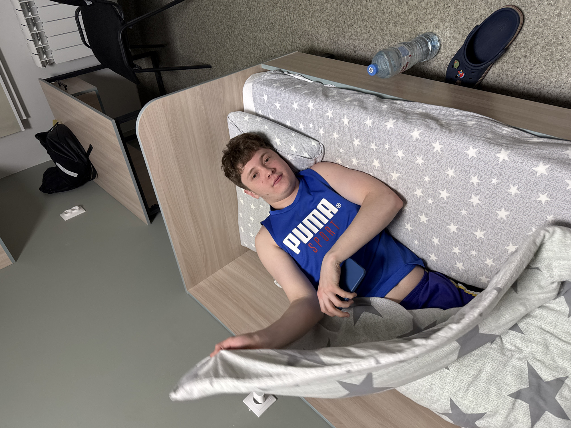 A young man lies in a bed with grey star-patterned sheets and a matching comforter. He wears a blue Puma tank top and blue shorts, holding a dark blue smartphone in his left hand. The bed has a wooden headboard. A water bottle and a dark blue Croc are on a shelf near the head of the bed.
In the background, a black chair and a backpack sit next to a desk with a light-colored wood grain finish. The floor is grey. A wall-mounted radiator is visible to the left.
The location is likely indoors, in Kazan, Russia.