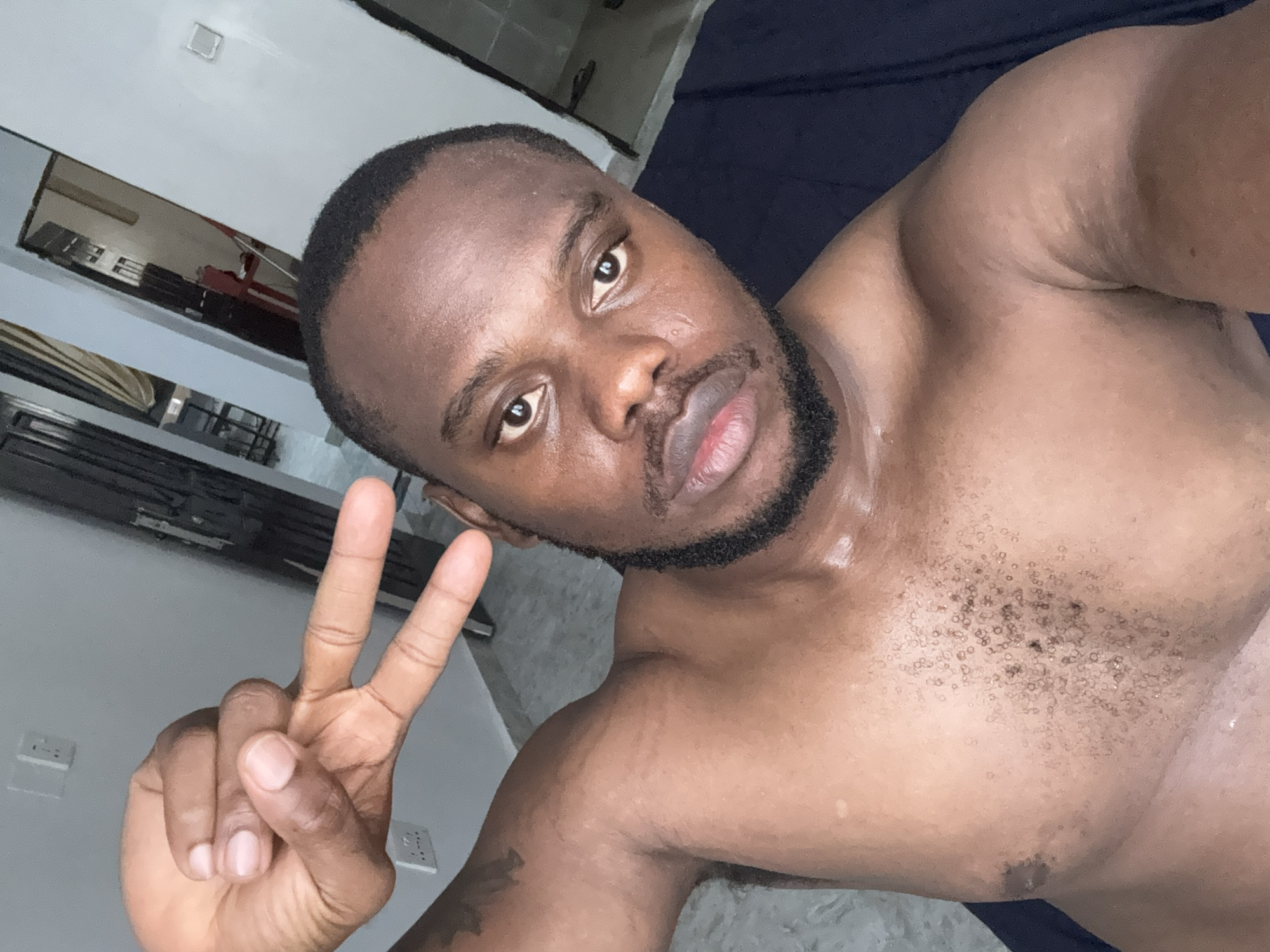 A shirtless, dark-skinned man with a shaved head and short beard is making a peace sign with his right hand. The man is indoors. The background shows walls, flooring, and a reflective surface possibly a mirror. The location of Iddo Tudunwada, Nigeria, cannot be verified from the image.